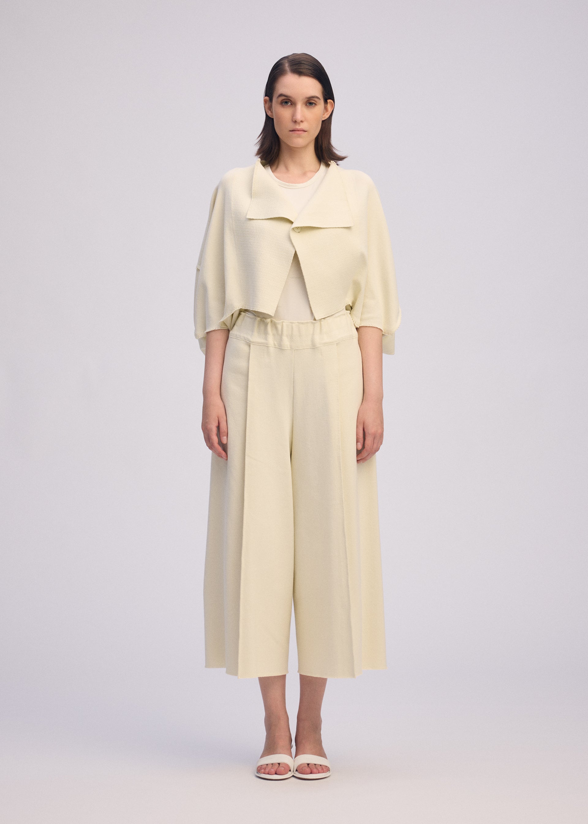 Zoomed view A woman stands facing forward against a plain light background, wearing the ISSEY MIYAKE CAMPAGNE cream cropped jacket and stretchy wide A-POC pants, paired with white sandals.