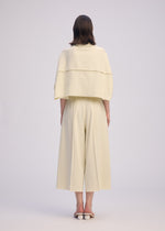 A person with straight, shoulder-length hair wears ISSEY MIYAKE's CAMPAGNE: a cream, loose top with a layered back and wide, stretchy A-POC pants, styled with white open-back shoes against a plain light background.