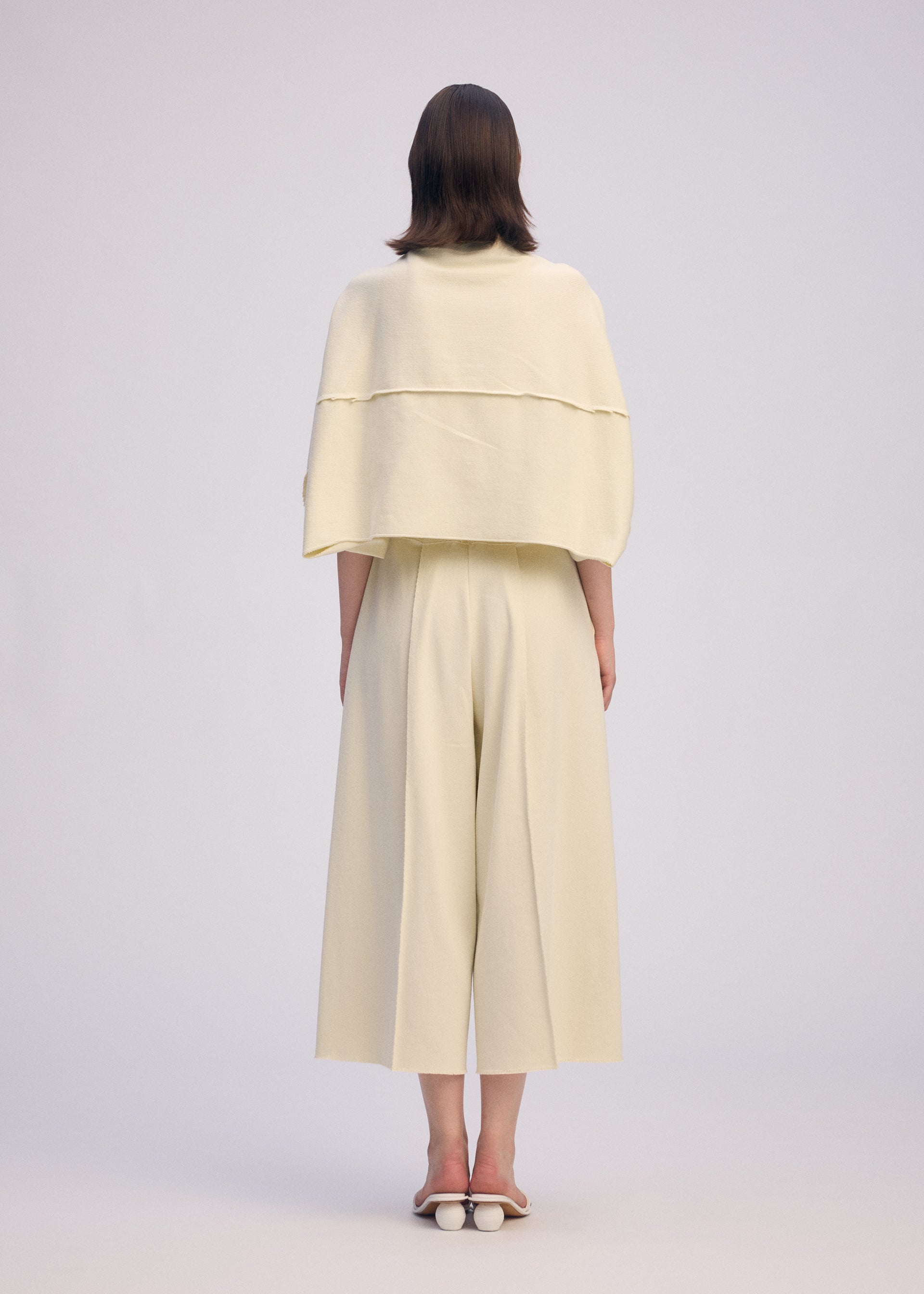 Zoomed view A person with straight, shoulder-length hair wears ISSEY MIYAKE's CAMPAGNE: a cream, loose top with a layered back and wide, stretchy A-POC pants, styled with white open-back shoes against a plain light background.