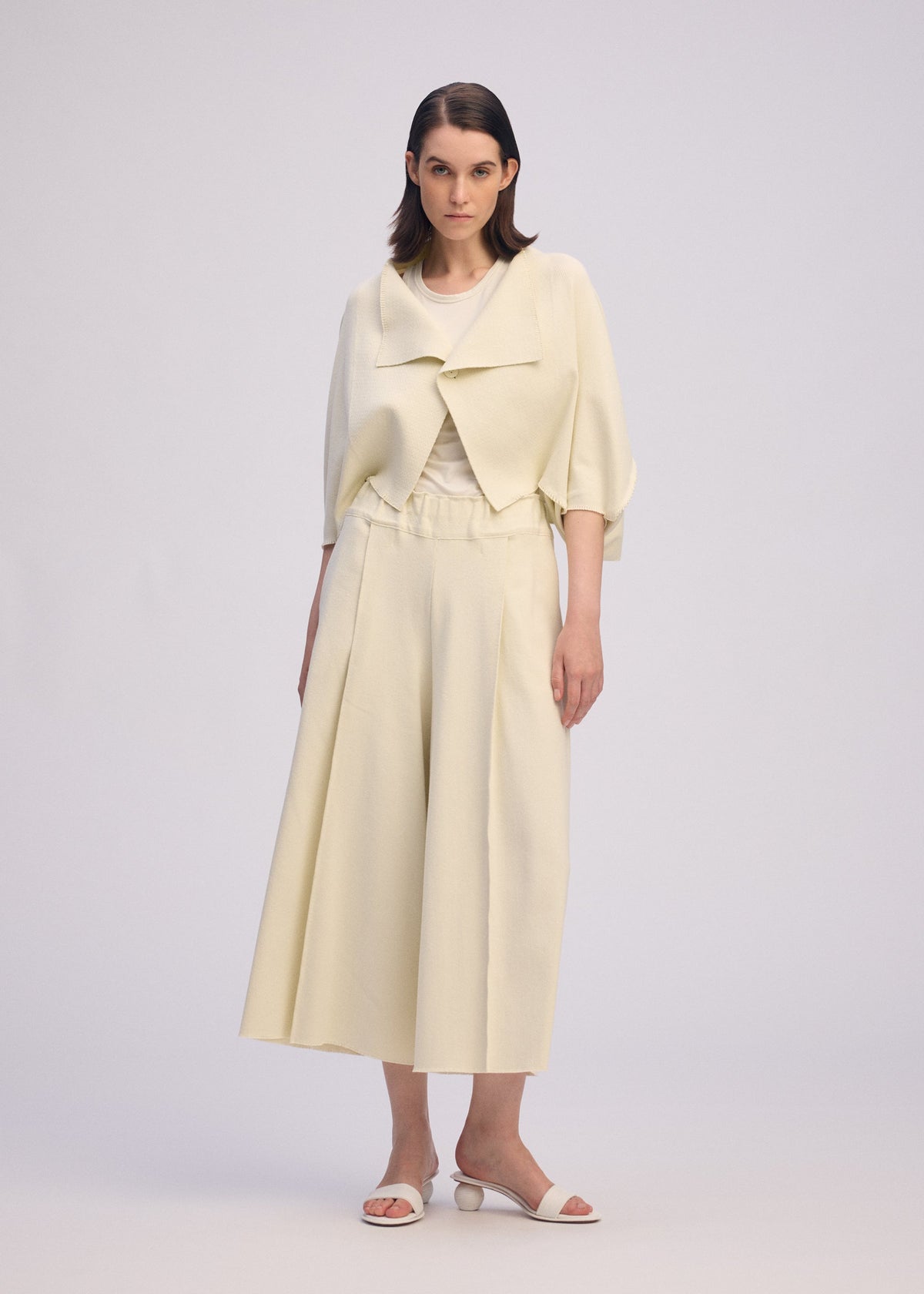 A woman stands facing forward in ISSEY MIYAKE's CAMPAGNE: a light cream outfit with a cropped jacket, stretchy wide pants, and white heeled sandals against a plain light background. view 5 of 6