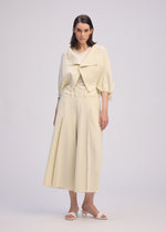 A woman stands facing forward in ISSEY MIYAKE's CAMPAGNE: a light cream outfit with a cropped jacket, stretchy wide pants, and white heeled sandals against a plain light background.