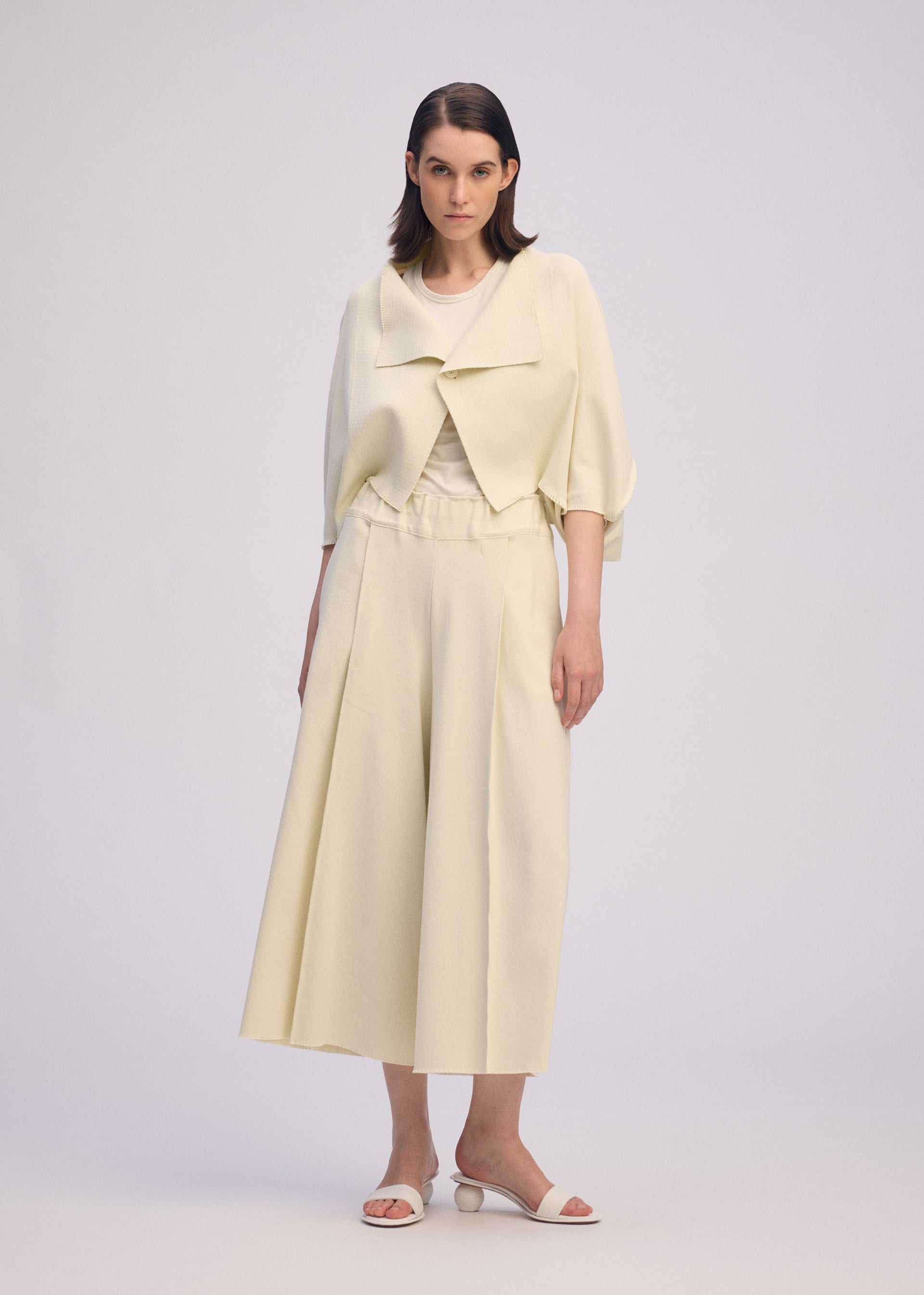 Zoomed view A woman stands facing forward in ISSEY MIYAKE's CAMPAGNE: a light cream outfit with a cropped jacket, stretchy wide pants, and white heeled sandals against a plain light background.