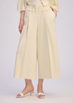A person wears ISSEY MIYAKE's CAMPAGNE wide-leg, high-waisted cream culotte pants made from stretchy A-POC material and cream open-toe heeled sandals, standing against a light background with hands in pockets; only the lower body is shown.