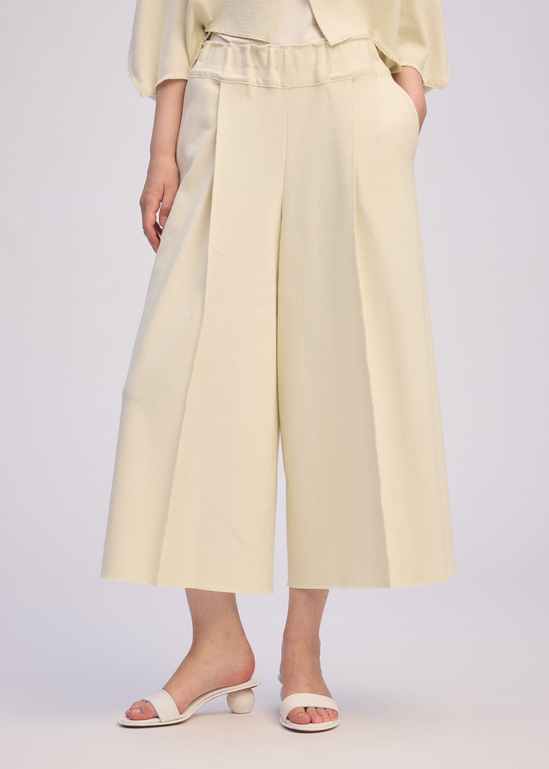 Zoomed view A person wears ISSEY MIYAKE's CAMPAGNE wide-leg, high-waisted cream culotte pants made from stretchy A-POC material and cream open-toe heeled sandals, standing against a light background with hands in pockets; only the lower body is shown.