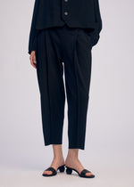 A person in CAMPAGNE black cropped pants by ISSEY MIYAKE, paired with a black buttoned top and open-toe heeled sandals, stands against a plain light background. The person's face is not shown.
