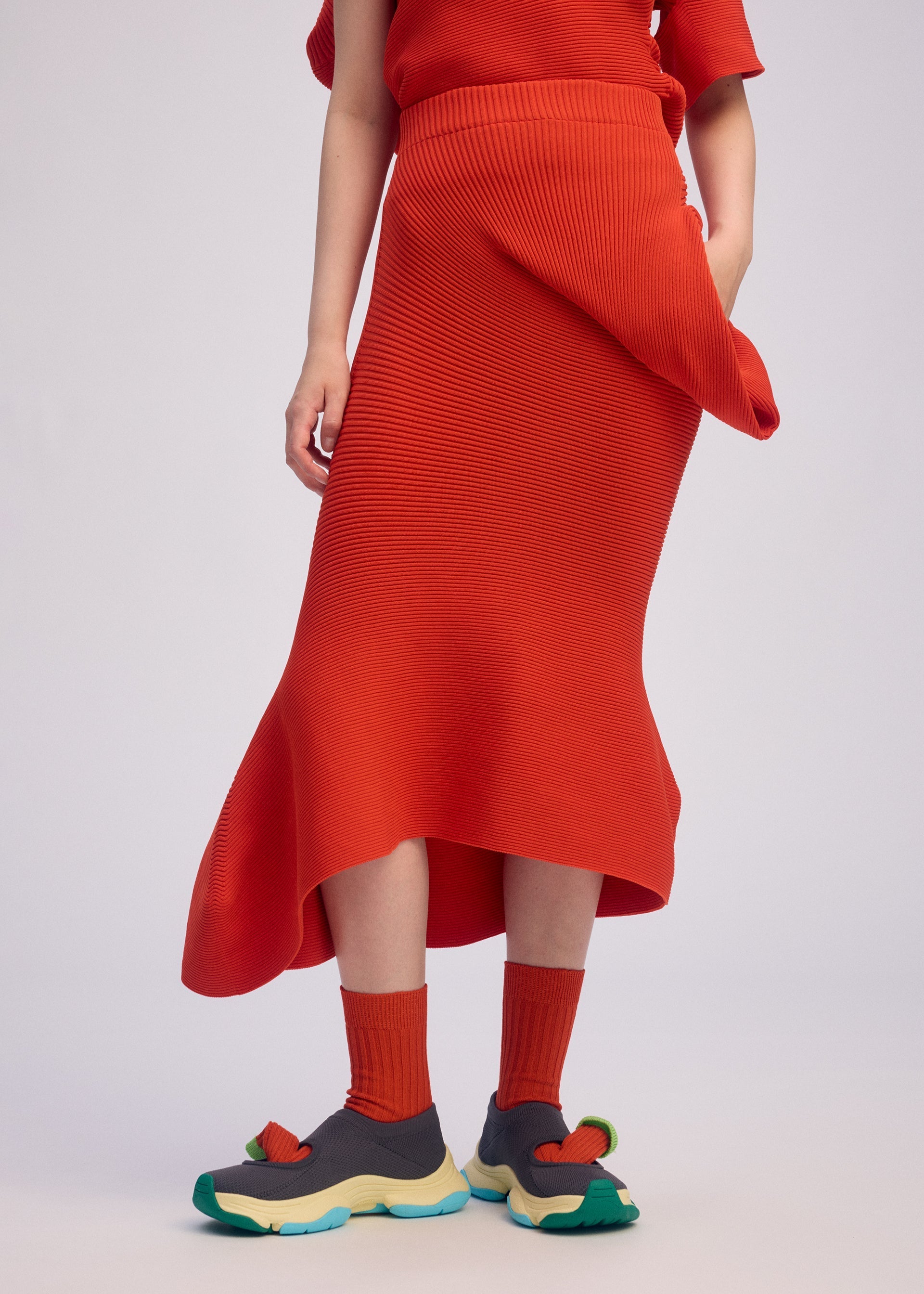 Zoomed view A person stands against a plain light background, wearing the ISSEY MIYAKE LIFE FORM textured red asymmetrical knit skirt made from recycled polyester, with matching socks and grey sneakers featuring colorful soles. The upper body is partially visible.