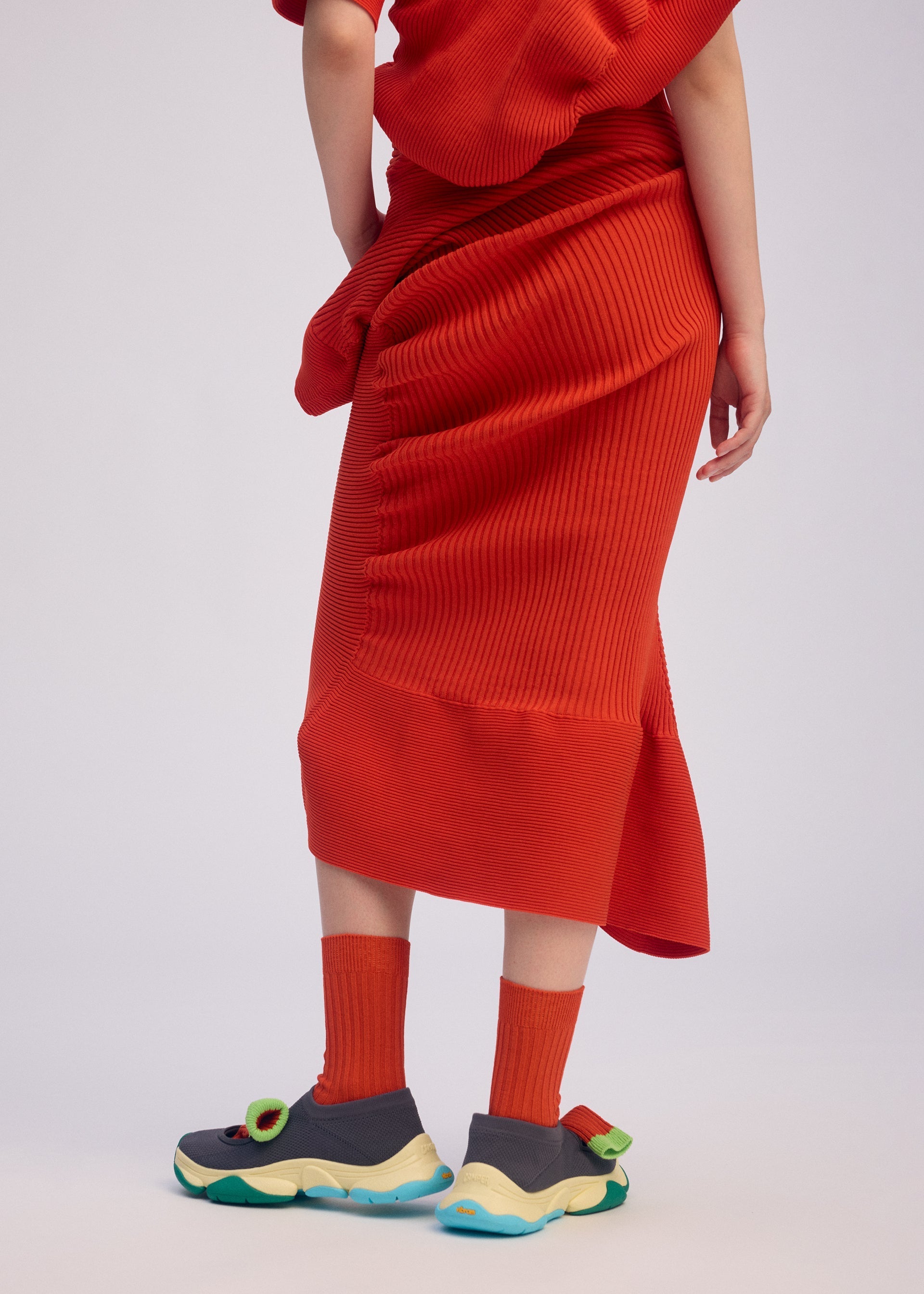 Zoomed view A person wears the LIFE FORM knit skirt and matching top by ISSEY MIYAKE, both crafted from recycled polyester, along with red socks and chunky gray-yellow sneakers with green accents. Their upper body is partially out of frame against a light background.