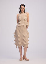 A woman with straight, shoulder-length hair and a neutral expression wears the ISSEY MIYAKE LINKAGE sleeveless beige dress with a unique silhouette and jagged hem, styled with open-toe sandals against a plain background.
