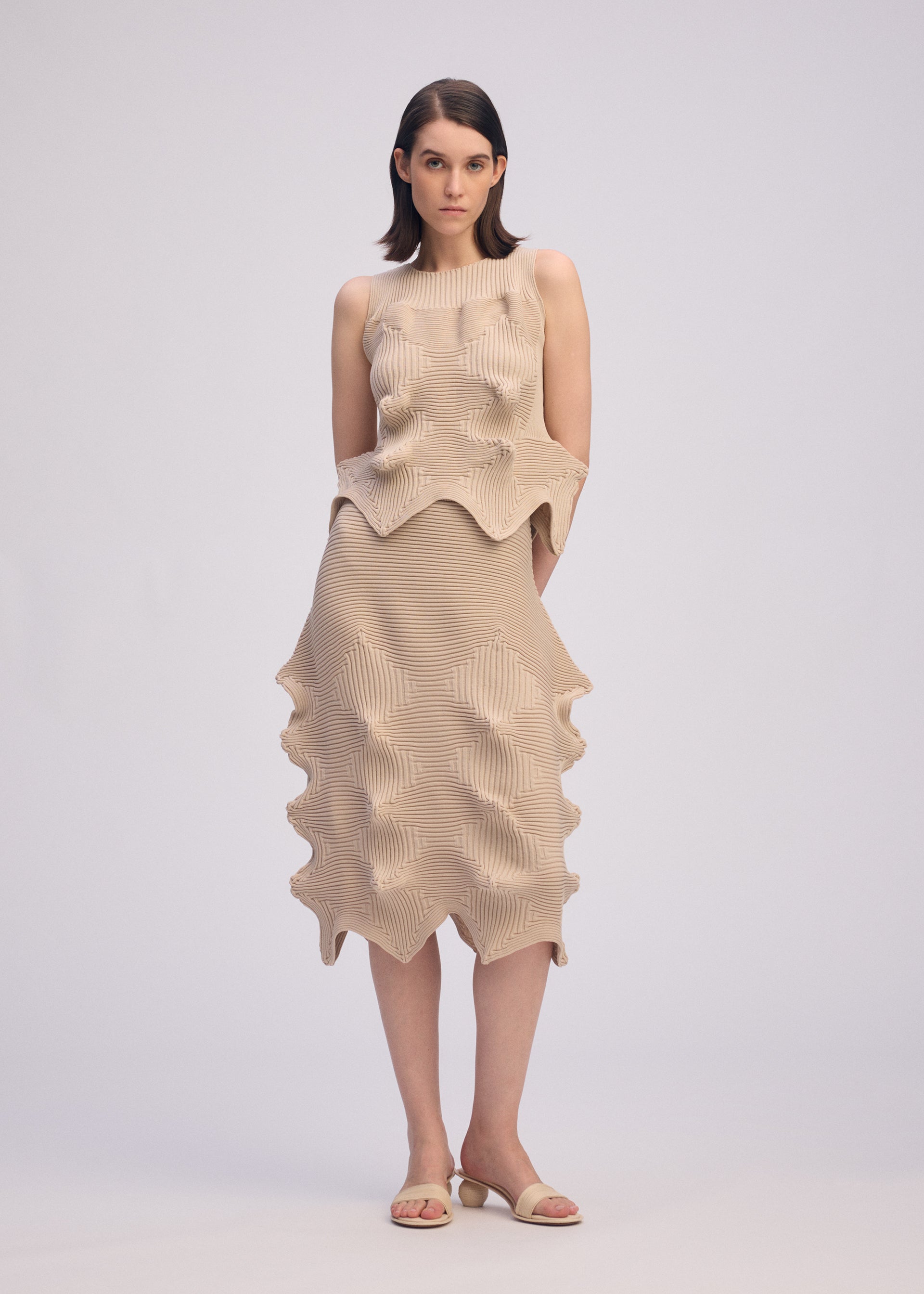 Zoomed view A woman with straight, shoulder-length hair and a neutral expression wears the ISSEY MIYAKE LINKAGE sleeveless beige dress with a unique silhouette and jagged hem, styled with open-toe sandals against a plain background.