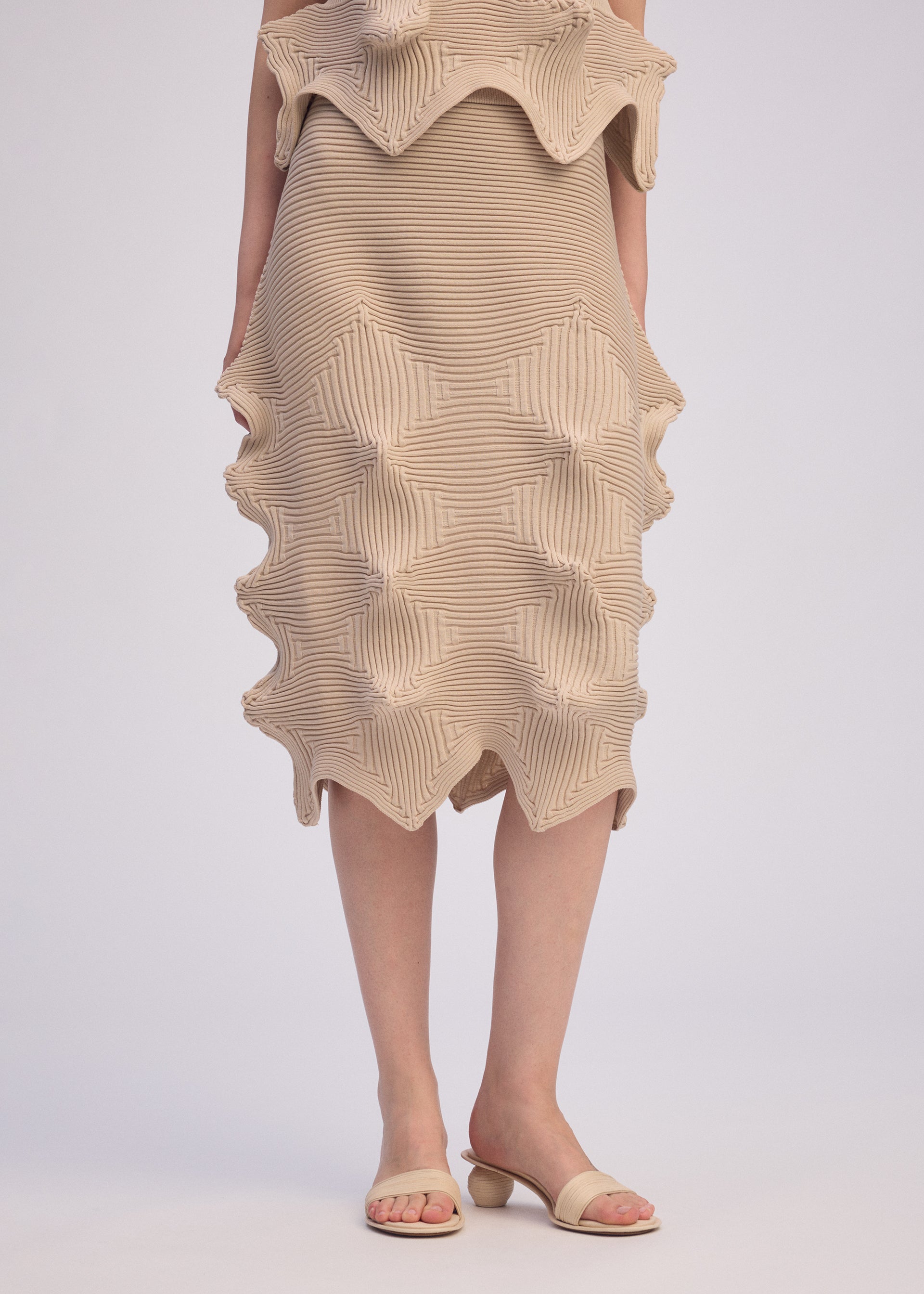 Zoomed view A person wears the ISSEY MIYAKE LINKAGE beige textured dress, crafted from recycled polyester with sculptural wavy details, paired with beige open-toe sandals with rounded heels. The image is cropped above the knees.
