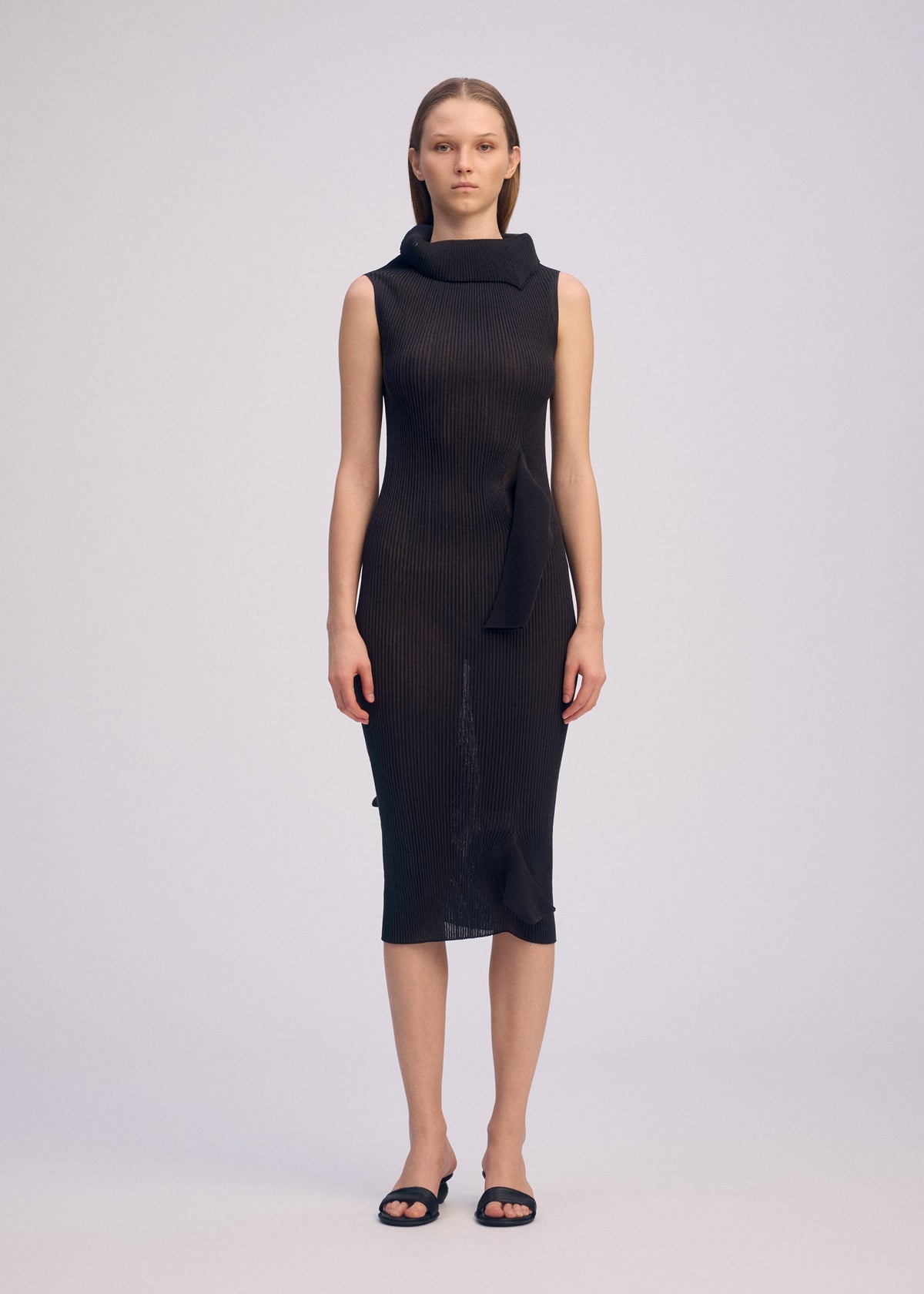 A woman with straight hair wears ISSEY MIYAKE’s ADVENTITIOUS：HIGH-NECKS sleeveless knit dress, featuring a high asymmetric collar and unique waist detailing, styled simply with black sandals against a plain light background. view 4 of 8