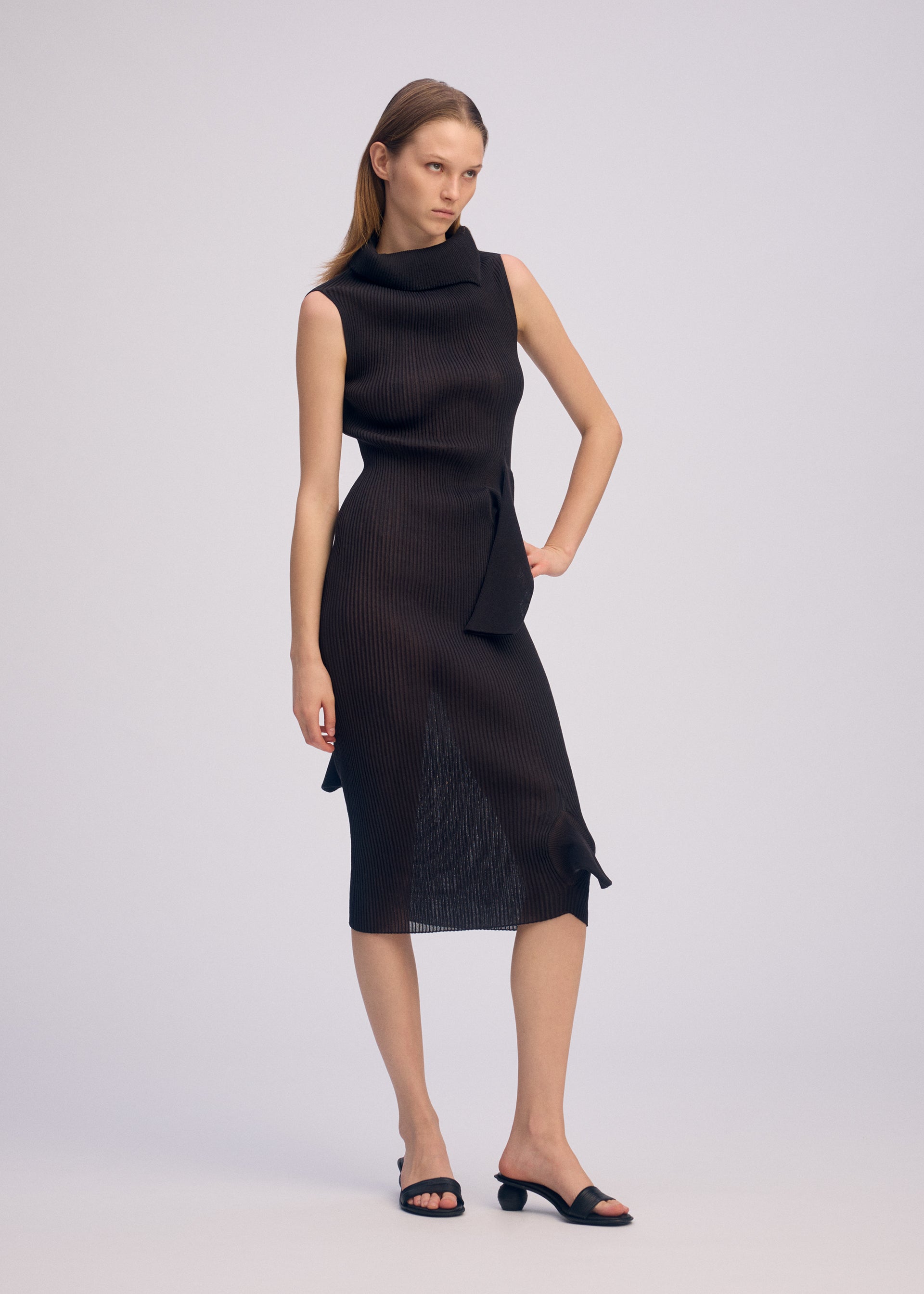 Zoomed view A woman poses confidently against a plain backdrop in ISSEY MIYAKE’s ADVENTITIOUS：HIGH-NECKS, a sleeveless textured black knit dress with a high neckline and subtle side accents, styled with black open-toe heeled sandals.
