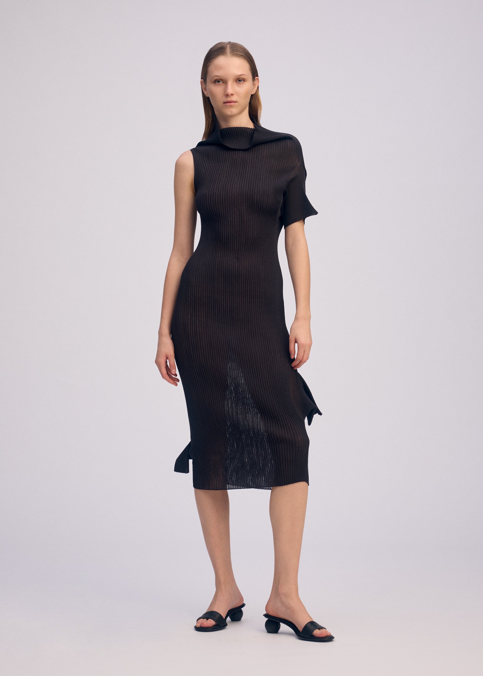 Zoomed view A woman poses before a plain backdrop in the ISSEY MIYAKE ADVENTITIOUS: HIGH-NECKS dress—a black, ribbed, sleeveless knit piece with an asymmetrical design and one short sleeve—paired with black open-toe low-heeled sandals.
