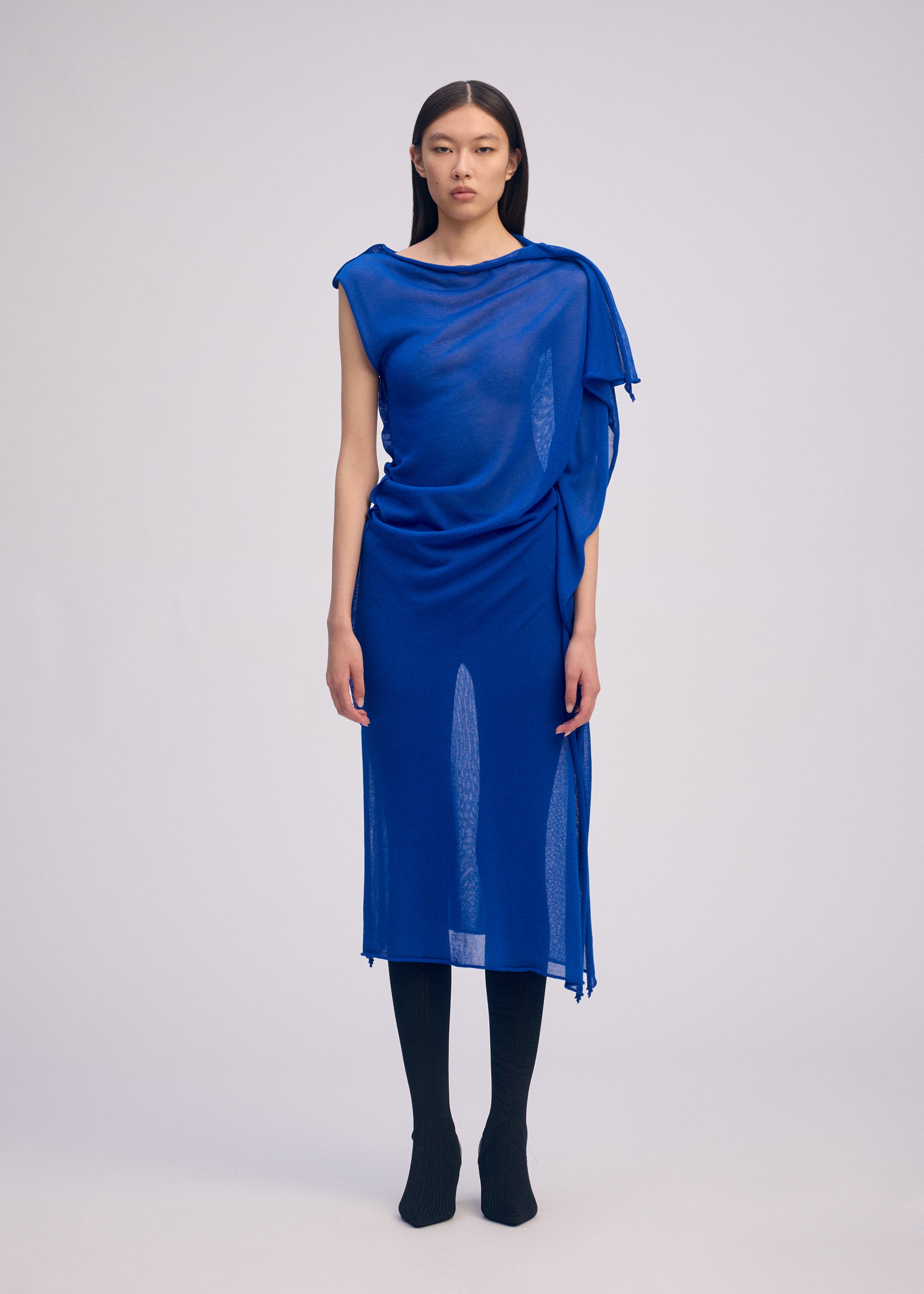 Zoomed view A person with straight dark hair wears the ISSEY MIYAKE CONCEALED KNIT, a sheer, asymmetrical royal blue lyocell blend dress with one shoulder exposed, paired with black tights and shoes against a plain light background.