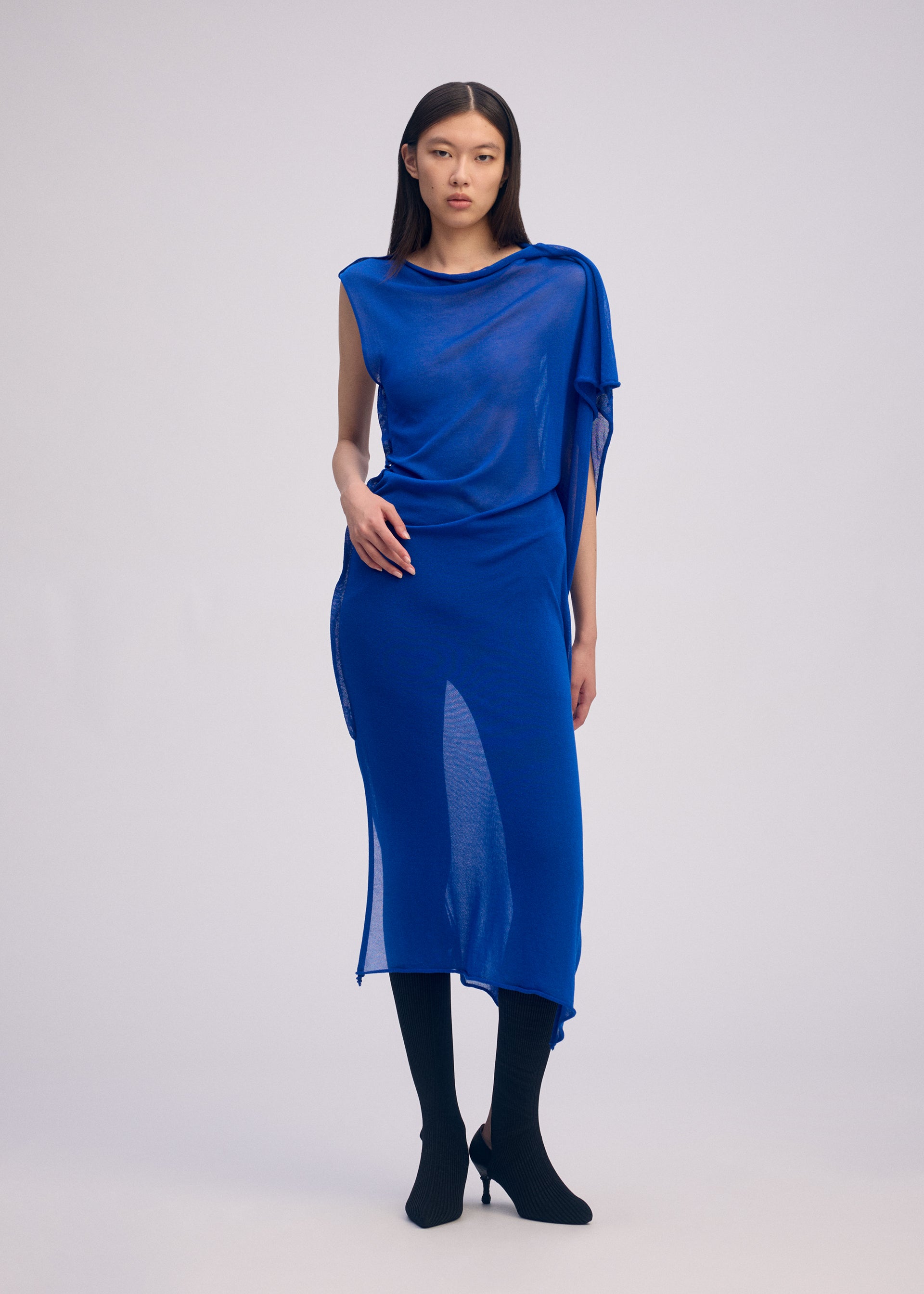 Zoomed view A woman with long dark hair stands against a light background, wearing ISSEY MIYAKE’s CONCEALED KNIT sheer asymmetrical bright blue lyocell blend dress over black tights and black heeled shoes, looking forward with a neutral expression.