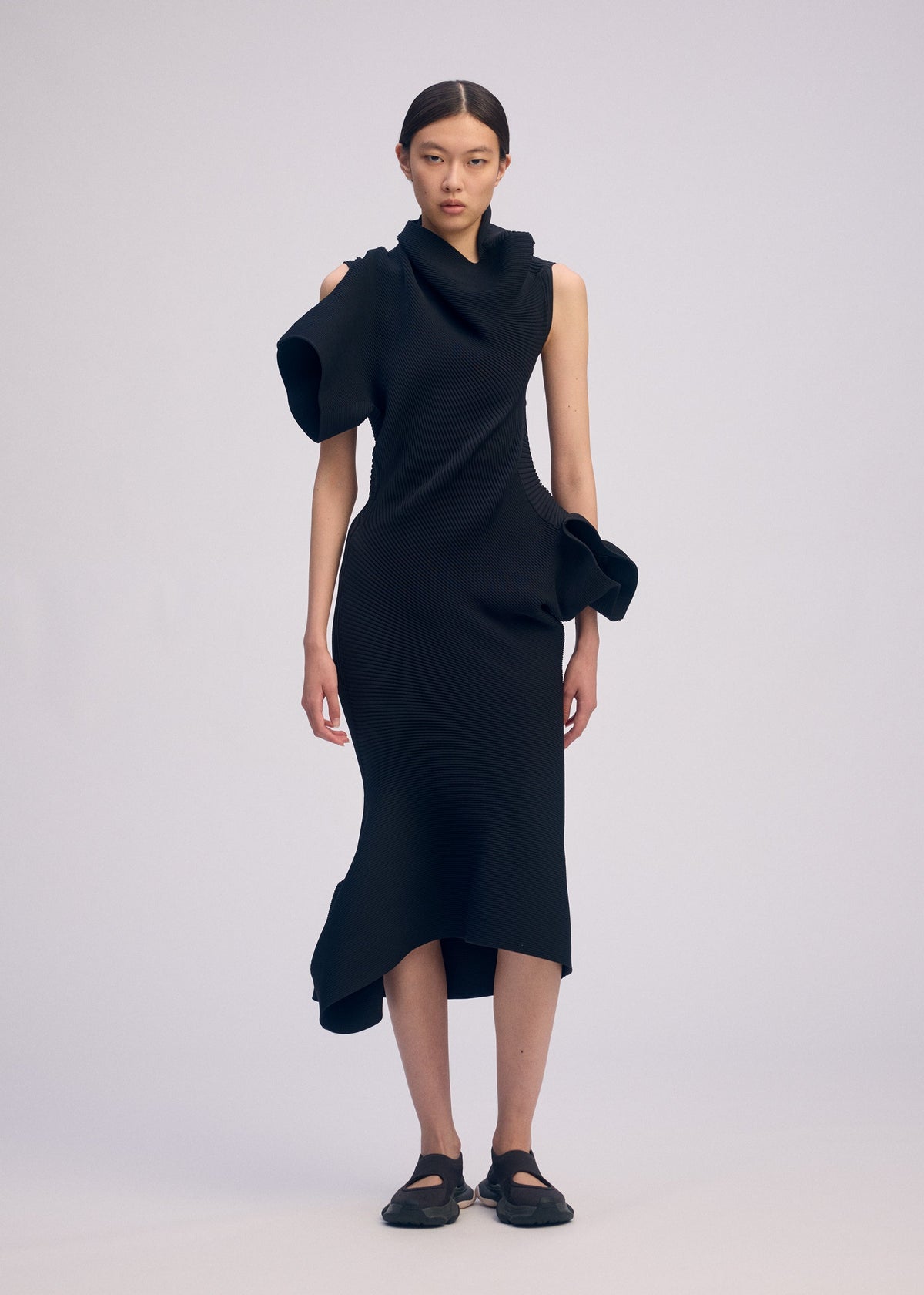 A person wears ISSEY MIYAKE’s LIFE FORM, a sleeveless asymmetric black knit dress crafted from recycled polyester with sculptural ruffles on shoulder and hip, paired with black shoes, against a plain light background for easy care style. view 5 of 7