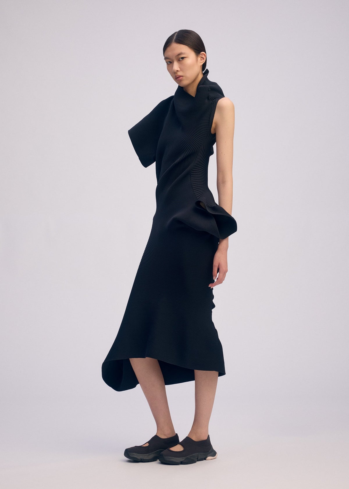 A woman in ISSEY MIYAKE’s LIFE FORM dress—an asymmetrical knit with sculptural ruffles, made from recycled polyester—and black shoes stands against a plain light background, her straight dark hair pulled back and her expression neutral. view 6 of 7