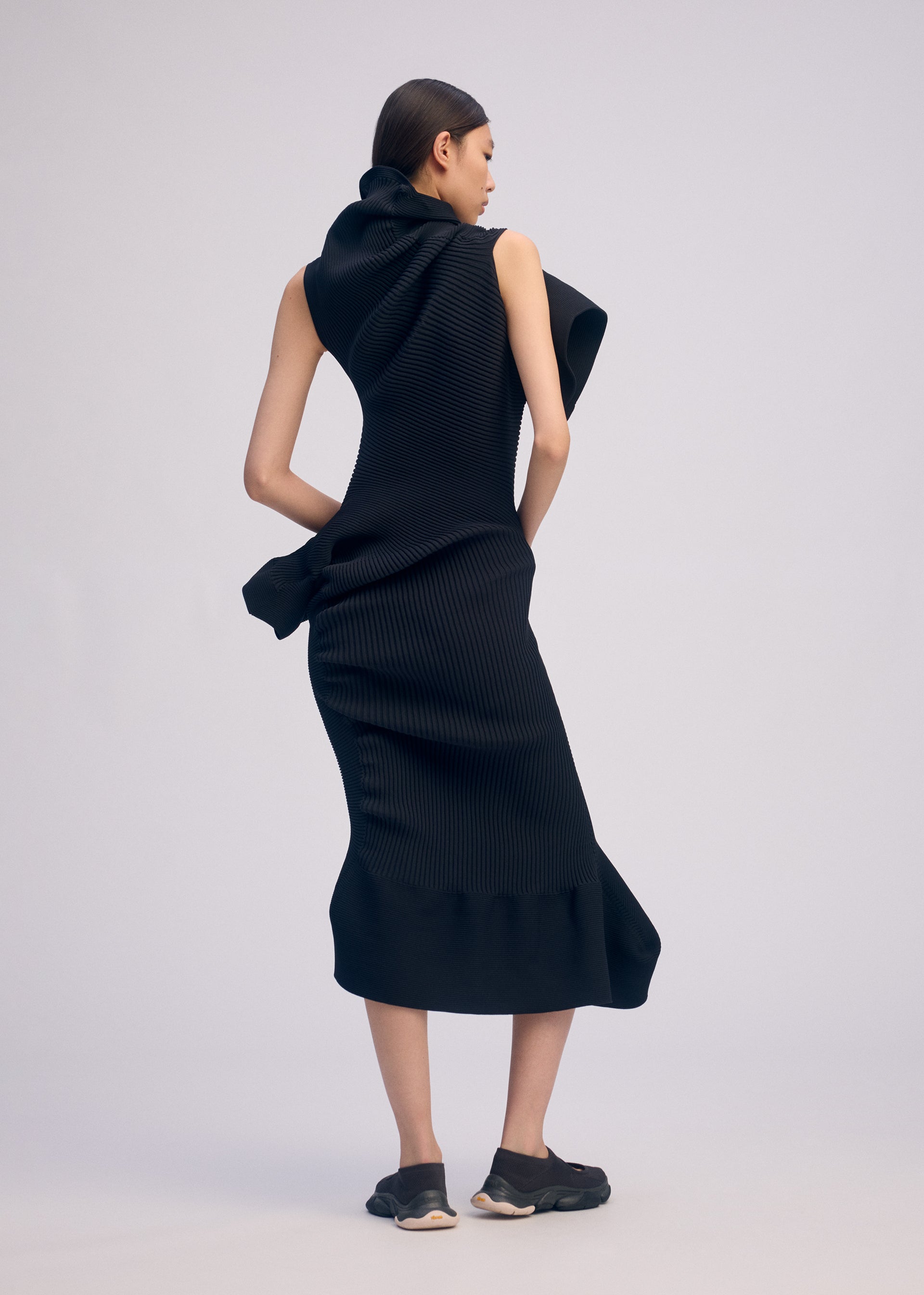 Zoomed view A woman with straight brown hair faces away, wearing the ISSEY MIYAKE LIFE FORM sleeveless black knit dress with sculptural folds, made from recycled polyester. She pairs it with black sneakers, set against a light background.