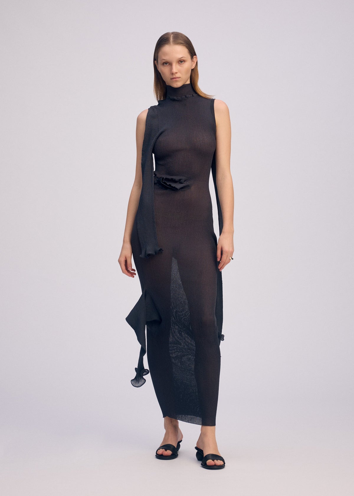 A woman models the ISSEY MIYAKE ADVENTITIOUS: SLEEVES sleeveless, sheer black knit dress with an asymmetrical hem and scarf-like detail against a plain background, styled with black open-toe sandals. view 7 of 8