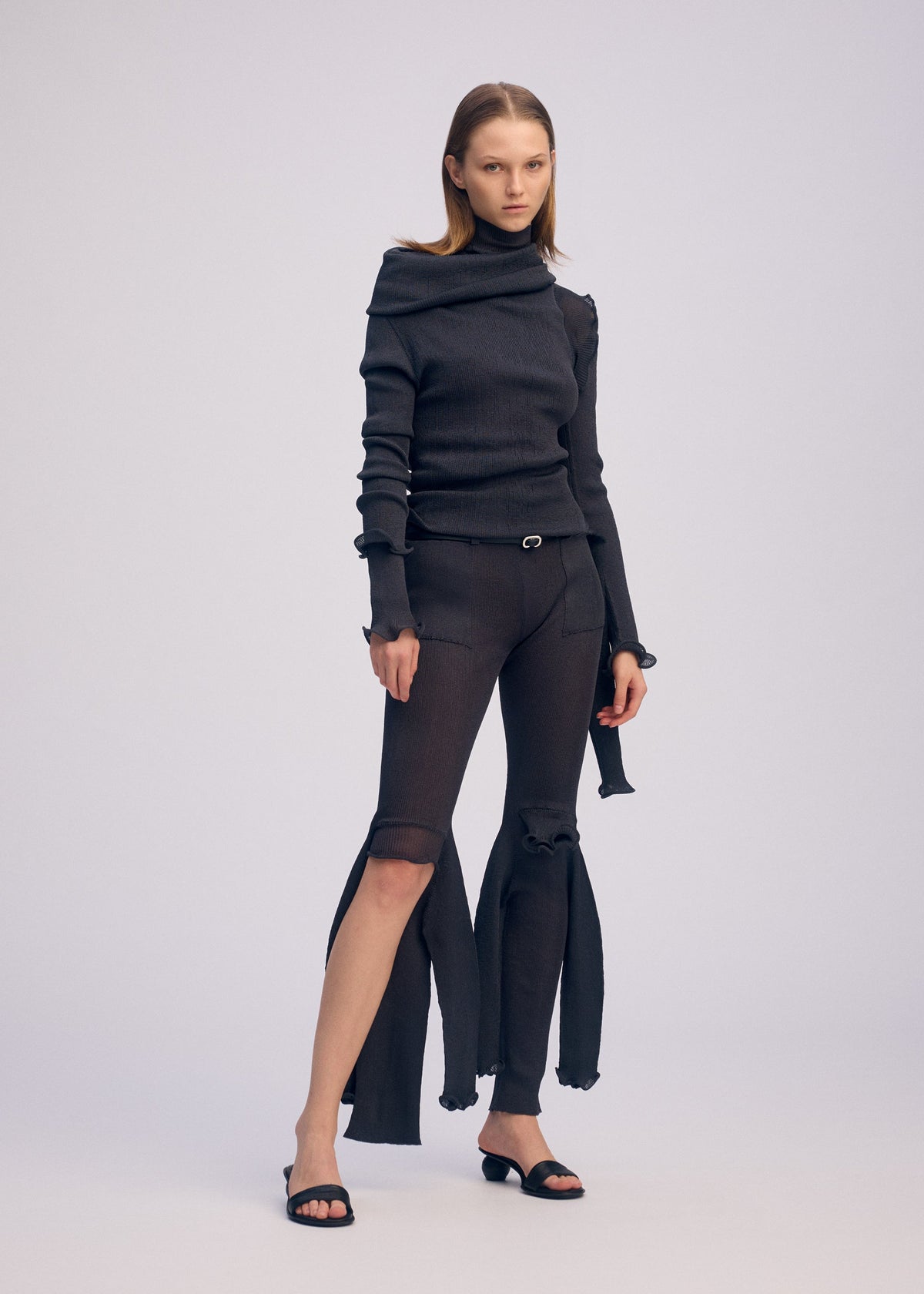 A woman models the ISSEY MIYAKE ADVENTITIOUS：SLEEVES—a fitted black turtleneck sweater—paired with striking black pants featuring open knee slits, black sandals, and a neutral expression against a plain background. view 8 of 8