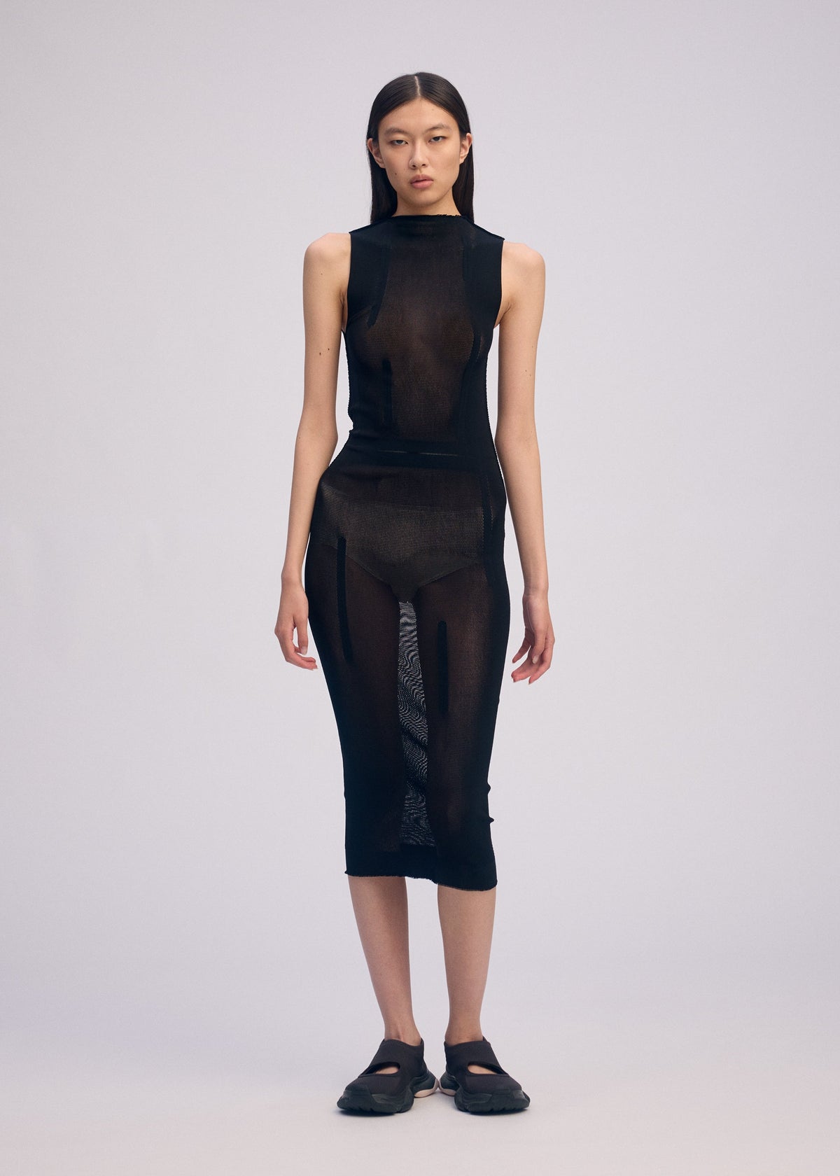 A woman with straight dark hair and a neutral expression wears the ISSEY MIYAKE SLITS MESH AP, a black semi-sheer mesh knitted midi dress with visible undergarments, paired with black closed-toe sandals against a plain background. view 4 of 5
