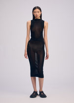 A woman with straight dark hair and a neutral expression wears the ISSEY MIYAKE SLITS MESH AP, a black semi-sheer mesh knitted midi dress with visible undergarments, paired with black closed-toe sandals against a plain background.