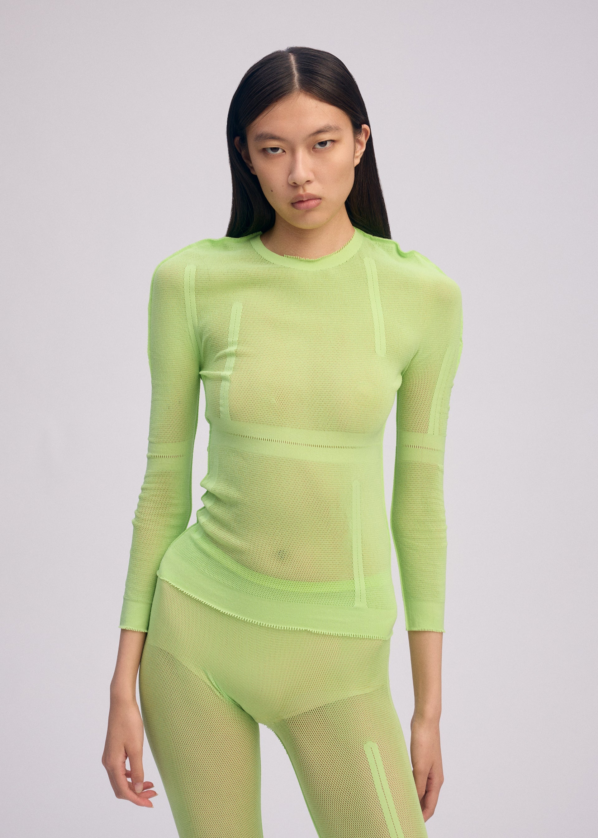 Zoomed view A person with long dark hair wears the ISSEY MIYAKE SLITS MESH AP, a sheer neon green knit top and pants perfect for layering, standing against a plain light background and gazing at the camera with a neutral expression.
