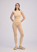 A person stands facing forward, wearing the ISSEY MIYAKE SLITS MESH AP beige sleeveless top over matching leggings and beige sneakers with yellow soles, set against a plain light gray background.