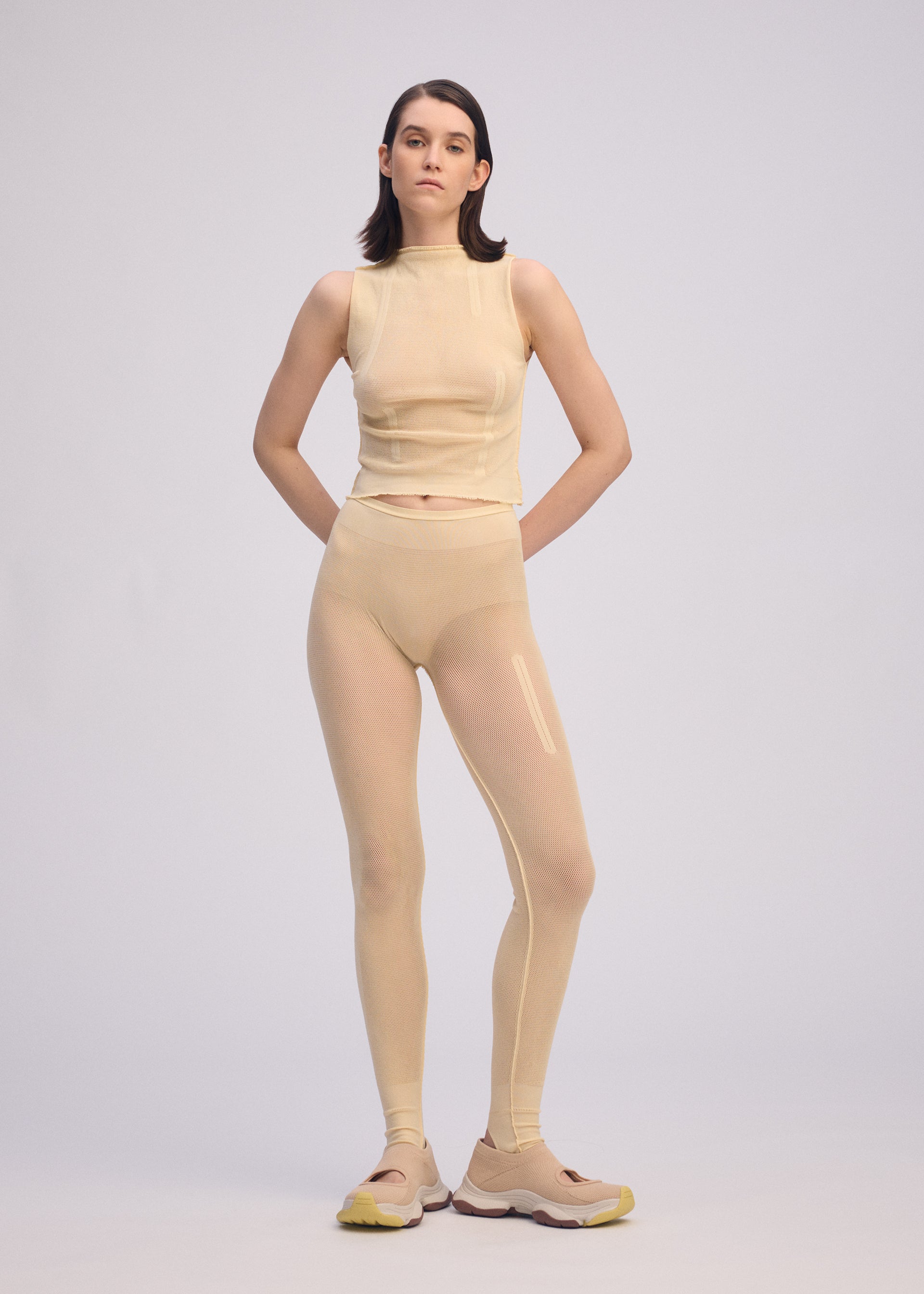 Zoomed view A person stands facing forward, wearing the ISSEY MIYAKE SLITS MESH AP beige sleeveless top over matching leggings and beige sneakers with yellow soles, set against a plain light gray background.