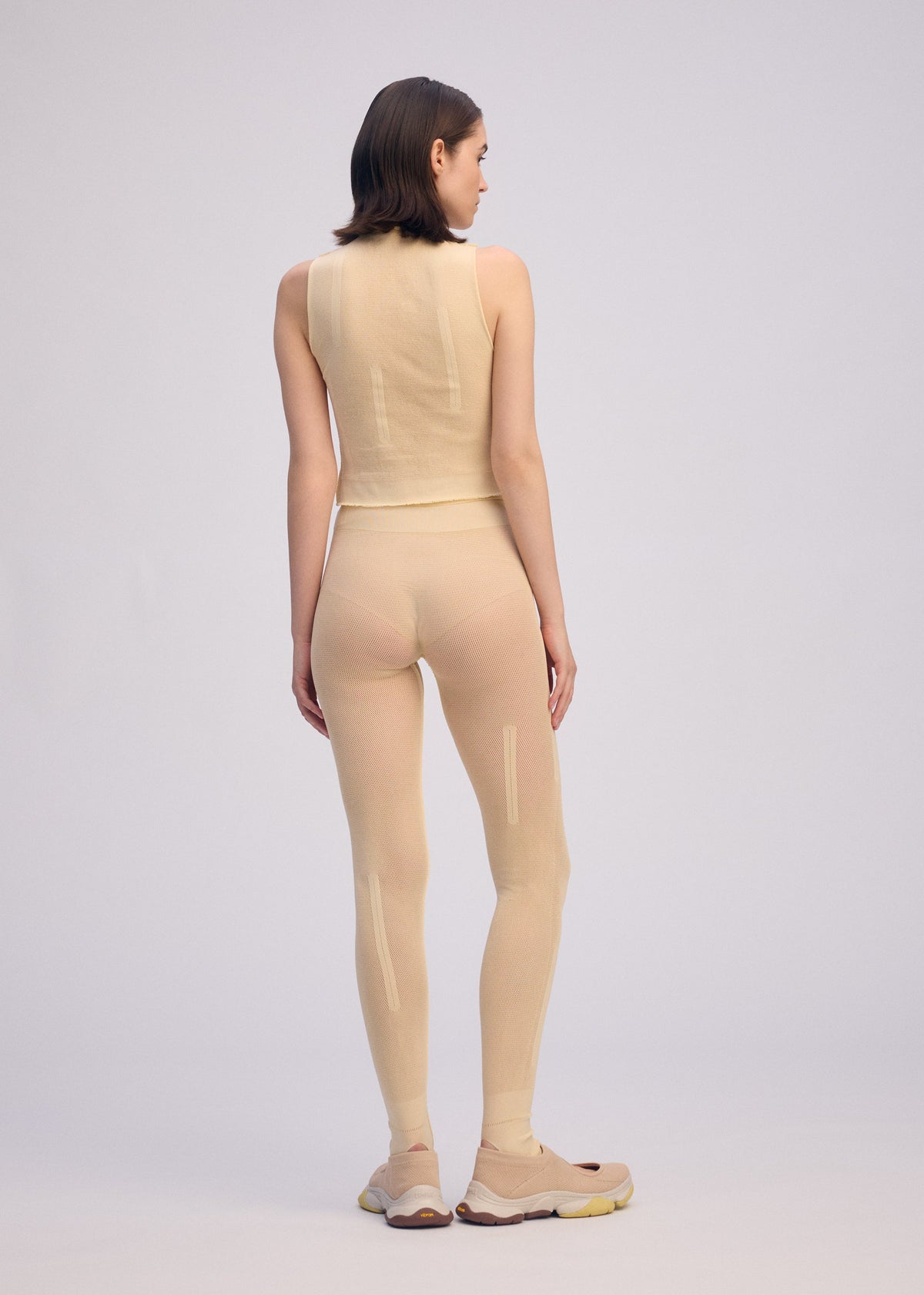 A person with short brown hair faces away, wearing ISSEY MIYAKE SLITS MESH AP—a fitted mesh sleeveless top and matching semi-sheer leggings as layered innerwear—with beige sneakers, set against a plain light background. view 5 of 6