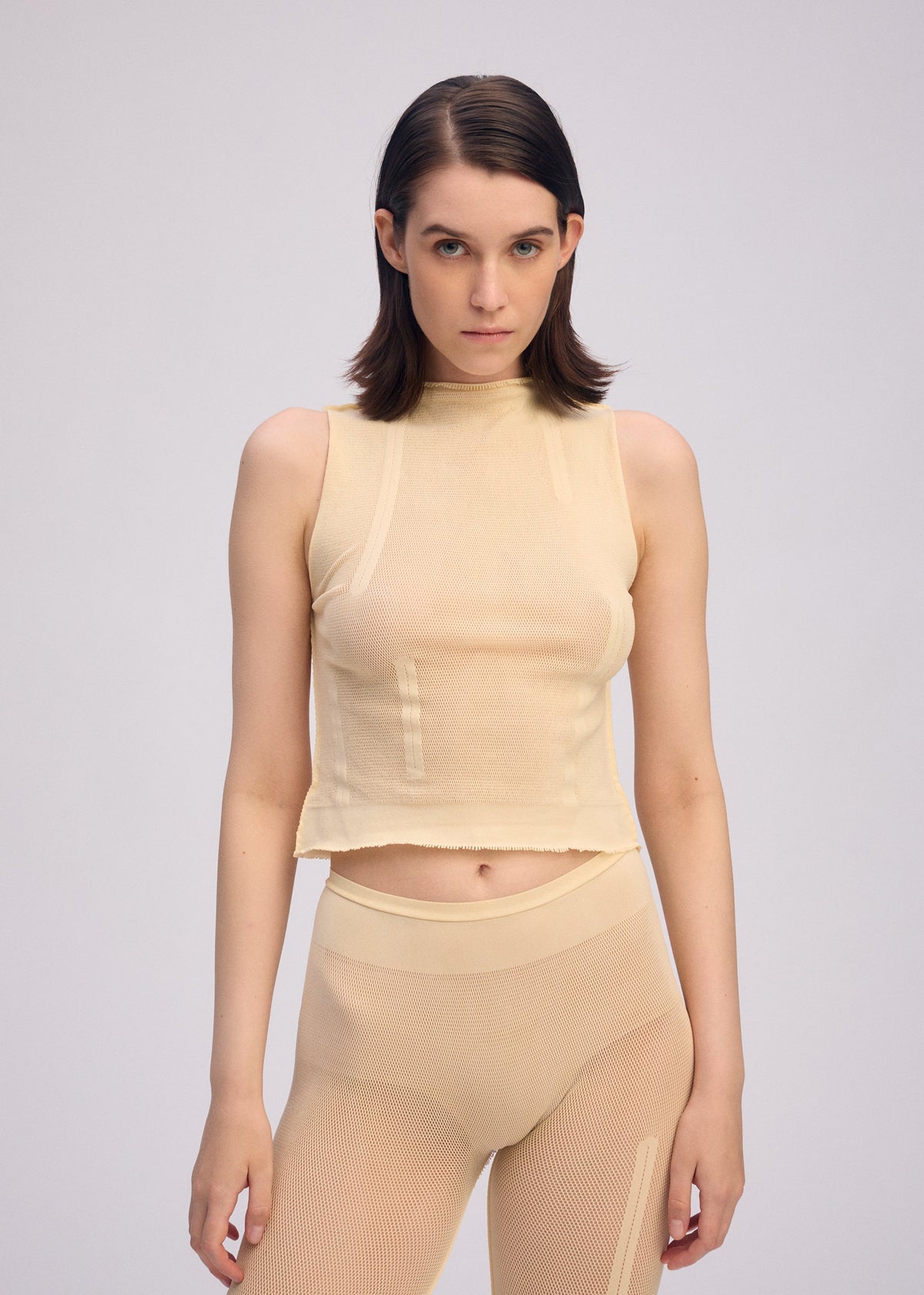 A person with straight, dark brown hair models the ISSEY MIYAKE SLITS MESH AP sleeveless top and matching high-waisted pants against a plain light gray background, looking directly at the camera with a neutral expression. view 6 of 6