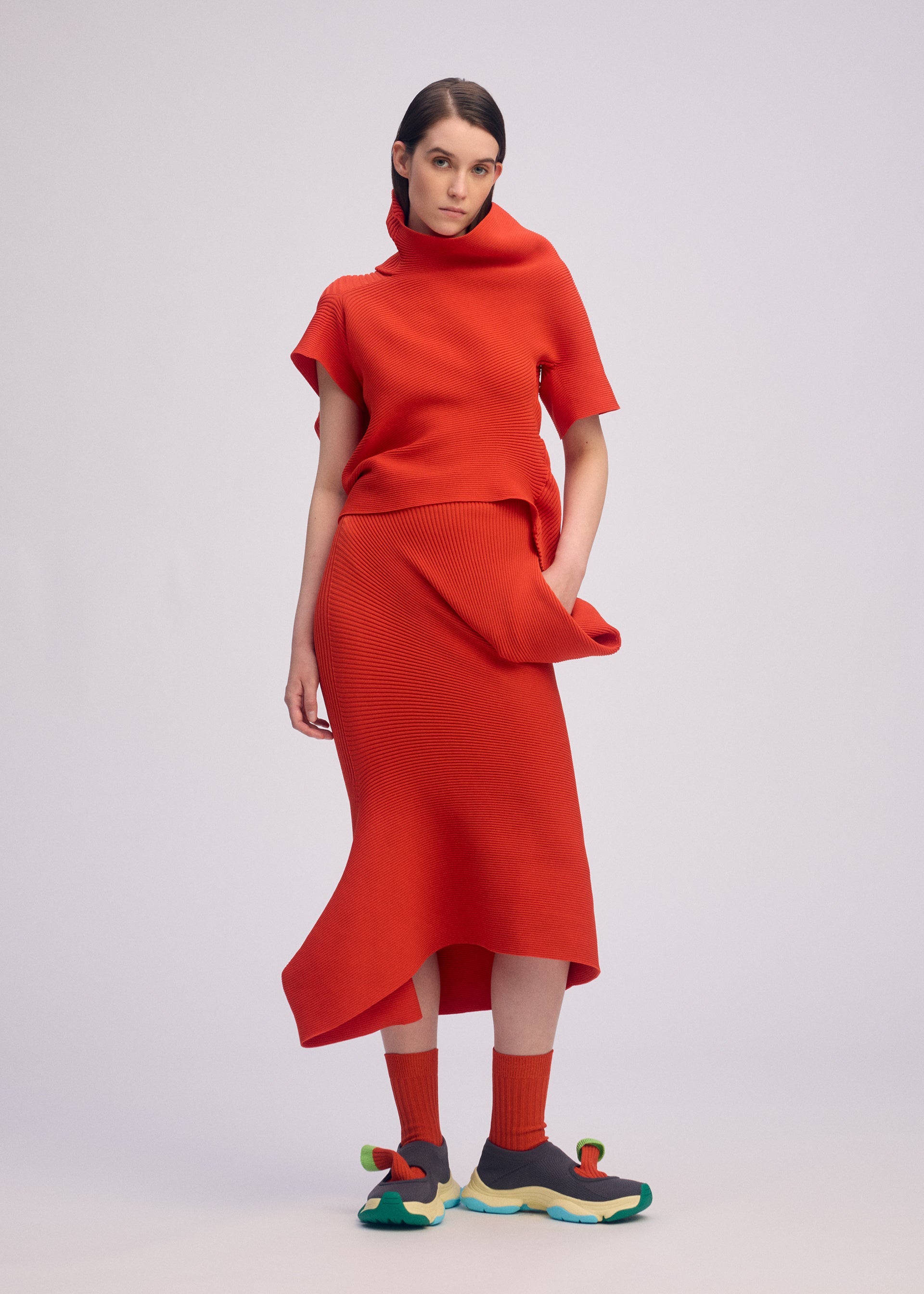 Zoomed view A woman poses against a light backdrop, wearing the LIFE FORM bright red asymmetrical knit top and skirt by ISSEY MIYAKE, made from recycled polyester. She pairs them with matching red socks and gray sneakers featuring yellow and green soles.