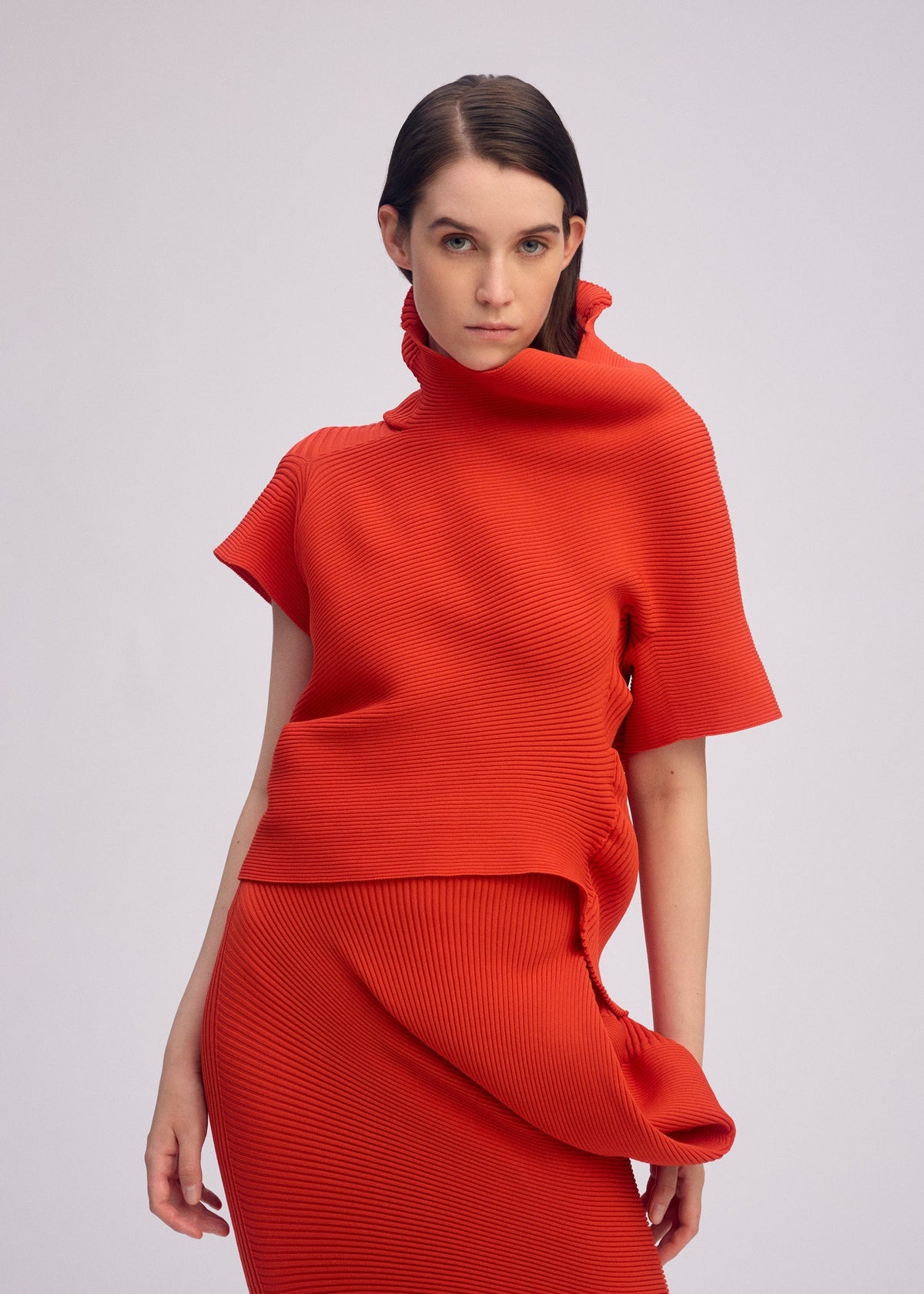 A woman with straight dark hair wears ISSEY MIYAKE’s LIFE FORM—a vivid red knit top with an asymmetrical high collar and matching skirt, both made from recycled polyester—standing against a plain background, gazing at the camera neutrally. view 6 of 7