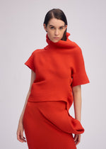 A woman with straight dark hair wears ISSEY MIYAKE’s LIFE FORM—a vivid red knit top with an asymmetrical high collar and matching skirt, both made from recycled polyester—standing against a plain background, gazing at the camera neutrally.