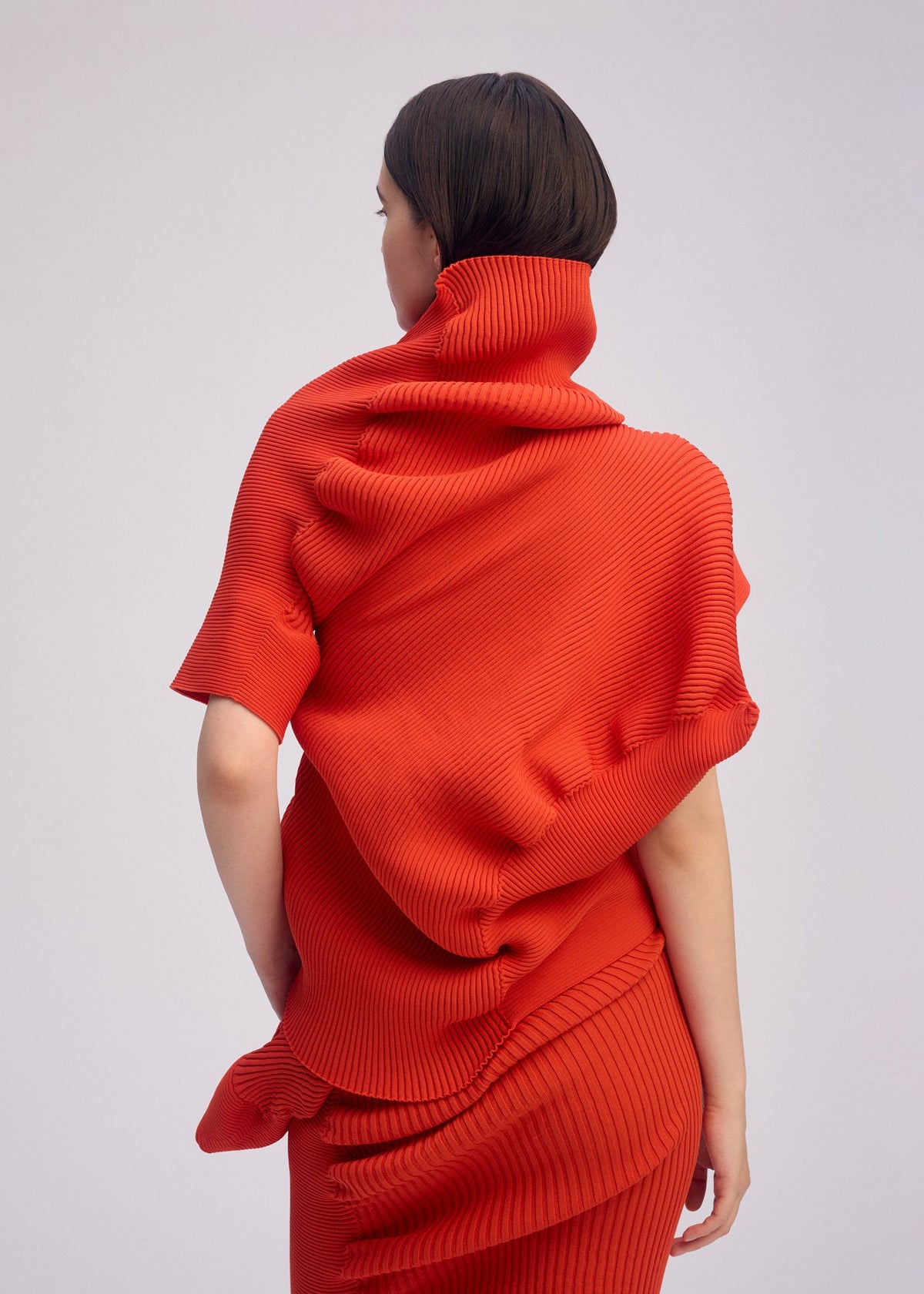A person with short dark hair is shown from behind, wearing ISSEY MIYAKE's LIFE FORM—a bright red, textured, sculptural outfit featuring pleats and organic folds—set against a plain light background. view 7 of 7