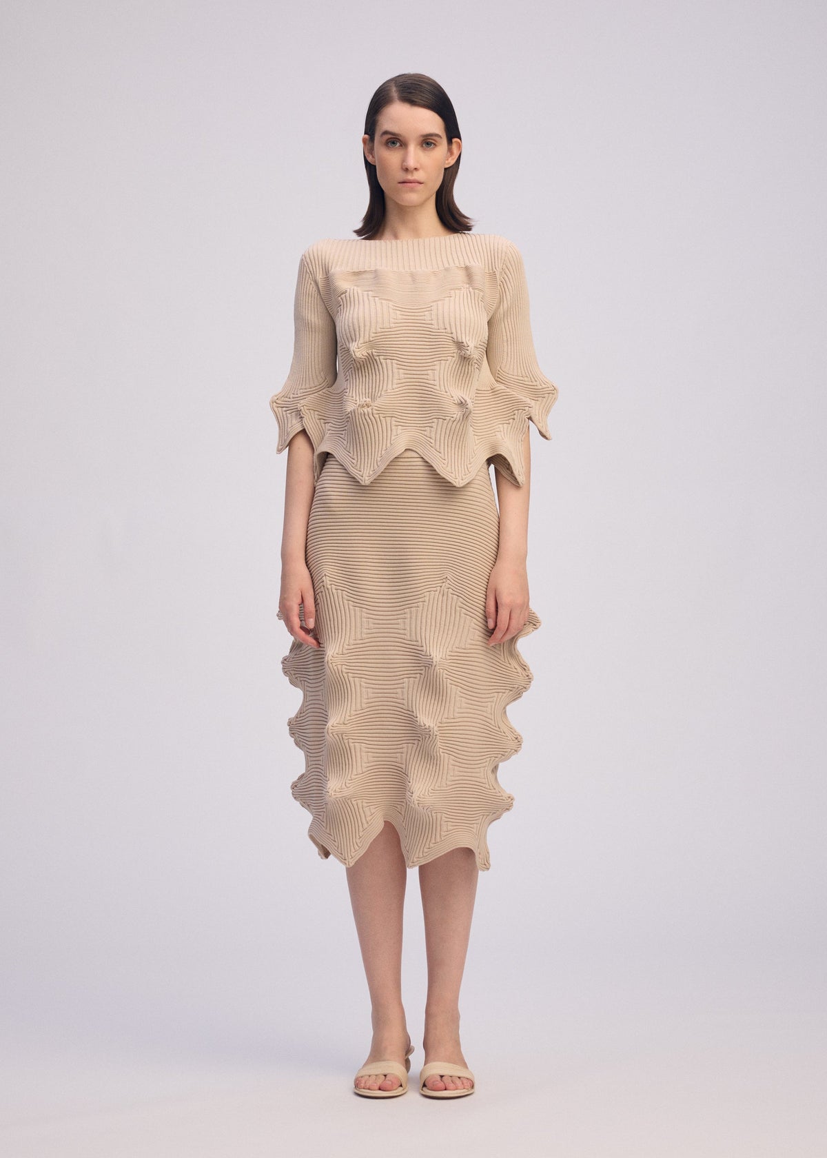 A woman faces forward in a unique silhouette wearing ISSEY MIYAKE's LINKAGE: a beige textured short-sleeved knit top and matching skirt with wavy edges. She has straight brown hair, beige sandals, and stands against a plain light background. view 3 of 5