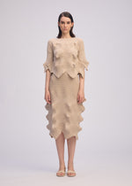 A woman faces forward in a unique silhouette wearing ISSEY MIYAKE's LINKAGE: a beige textured short-sleeved knit top and matching skirt with wavy edges. She has straight brown hair, beige sandals, and stands against a plain light background.