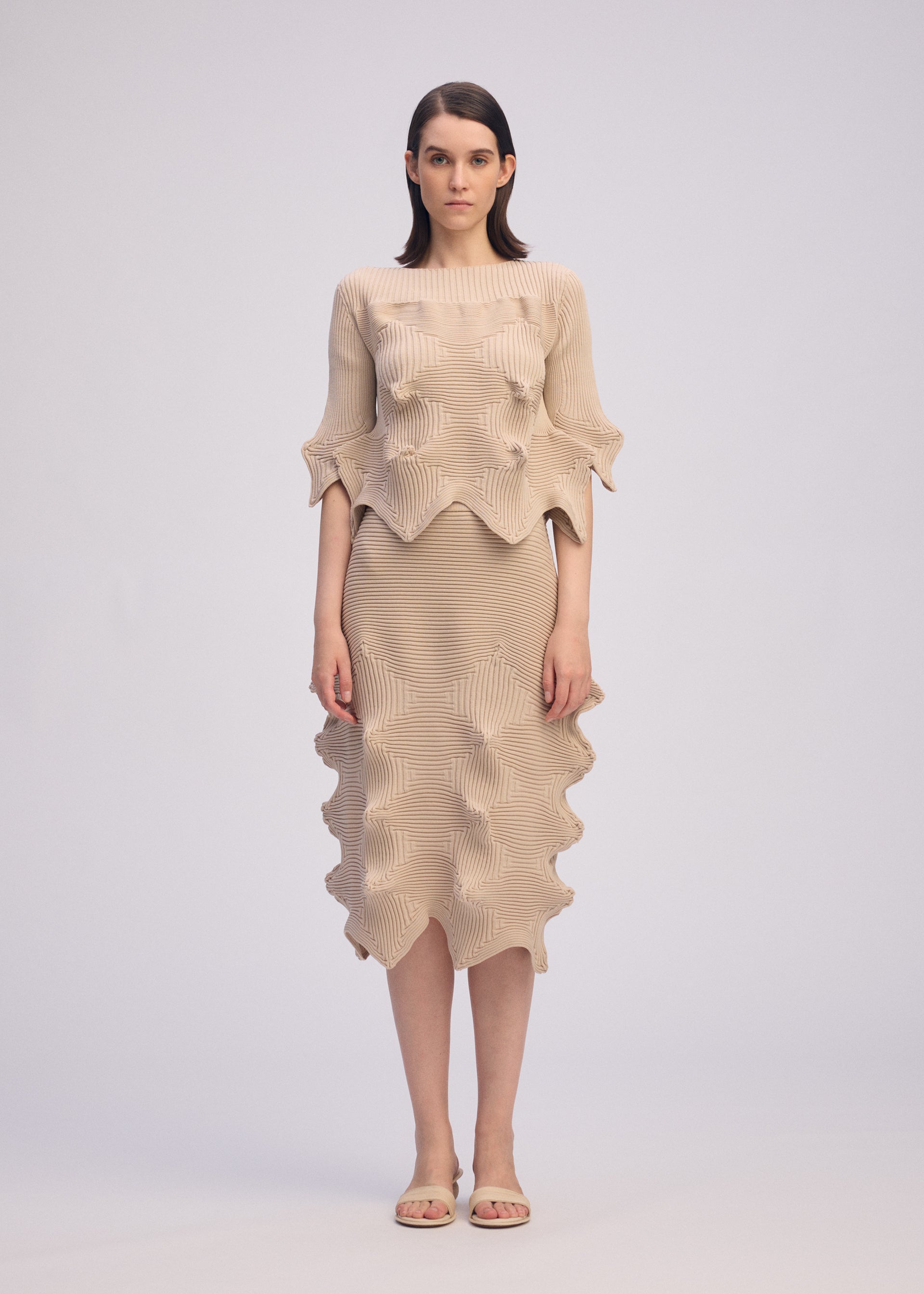 Zoomed view A woman faces forward in a unique silhouette wearing ISSEY MIYAKE's LINKAGE: a beige textured short-sleeved knit top and matching skirt with wavy edges. She has straight brown hair, beige sandals, and stands against a plain light background.