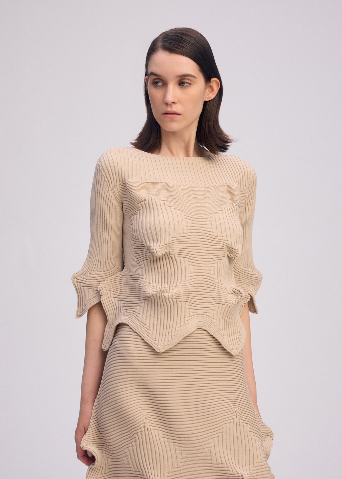 A woman with straight brown hair wears the ISSEY MIYAKE LINKAGE short-sleeved knit top and textured beige ribbed piece featuring sculpted star-like patterns, standing against a plain light background, looking slightly to the right. view 4 of 5
