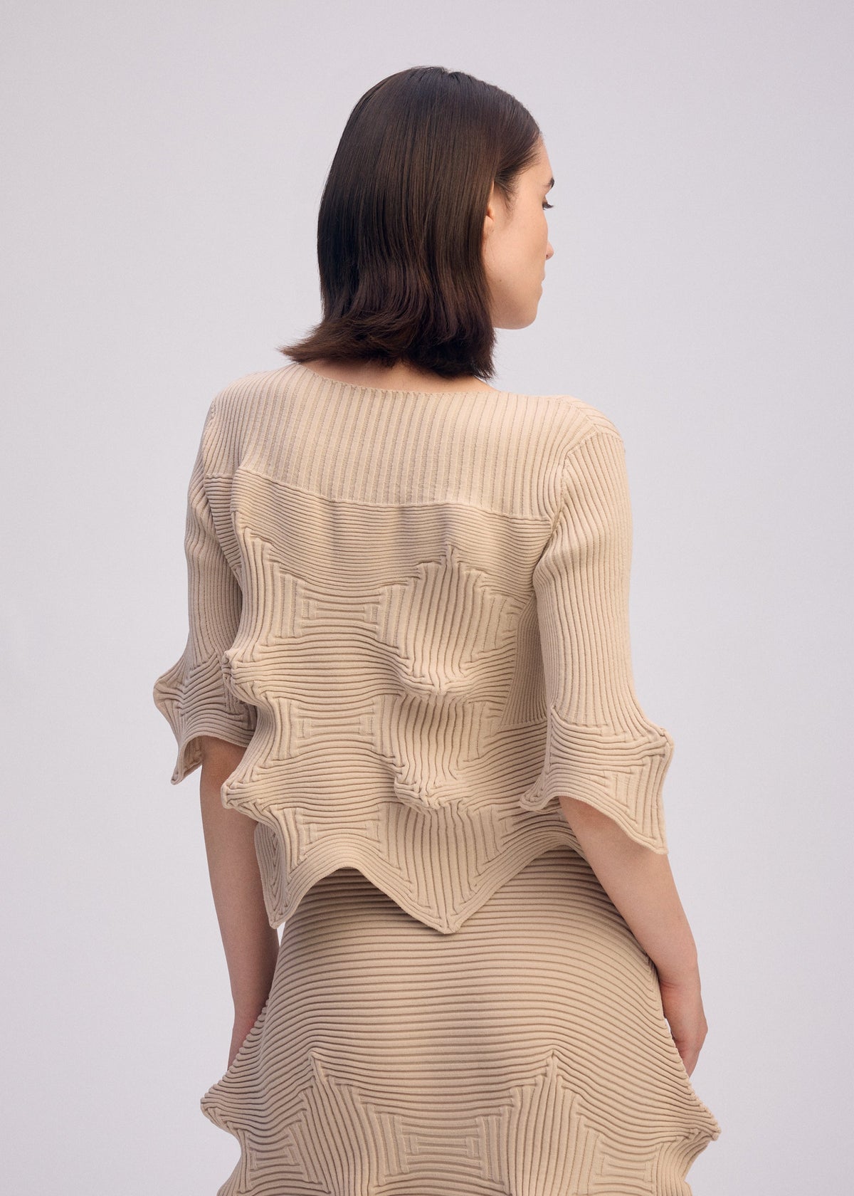 A woman with shoulder-length brown hair models ISSEY MIYAKE's LINKAGE set in beige recycled polyester, featuring sculptural, star-shaped details and short sleeves, against a plain light background. view 5 of 5