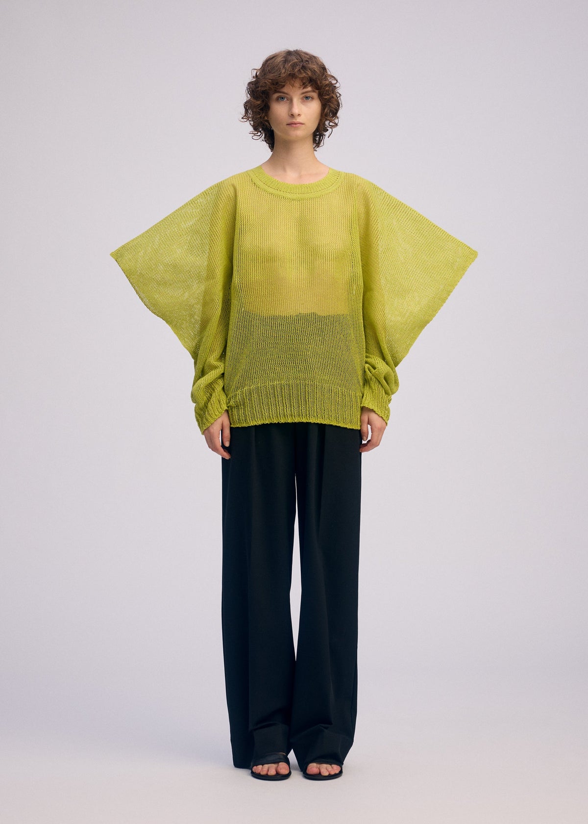 A person with short curly hair models the ISSEY MIYAKE CIALDA mesh-like yellow-green knit top layered over a yellow shirt, paired with loose black trousers and sandals, forming a voluminous silhouette against a plain light background. view 5 of 10