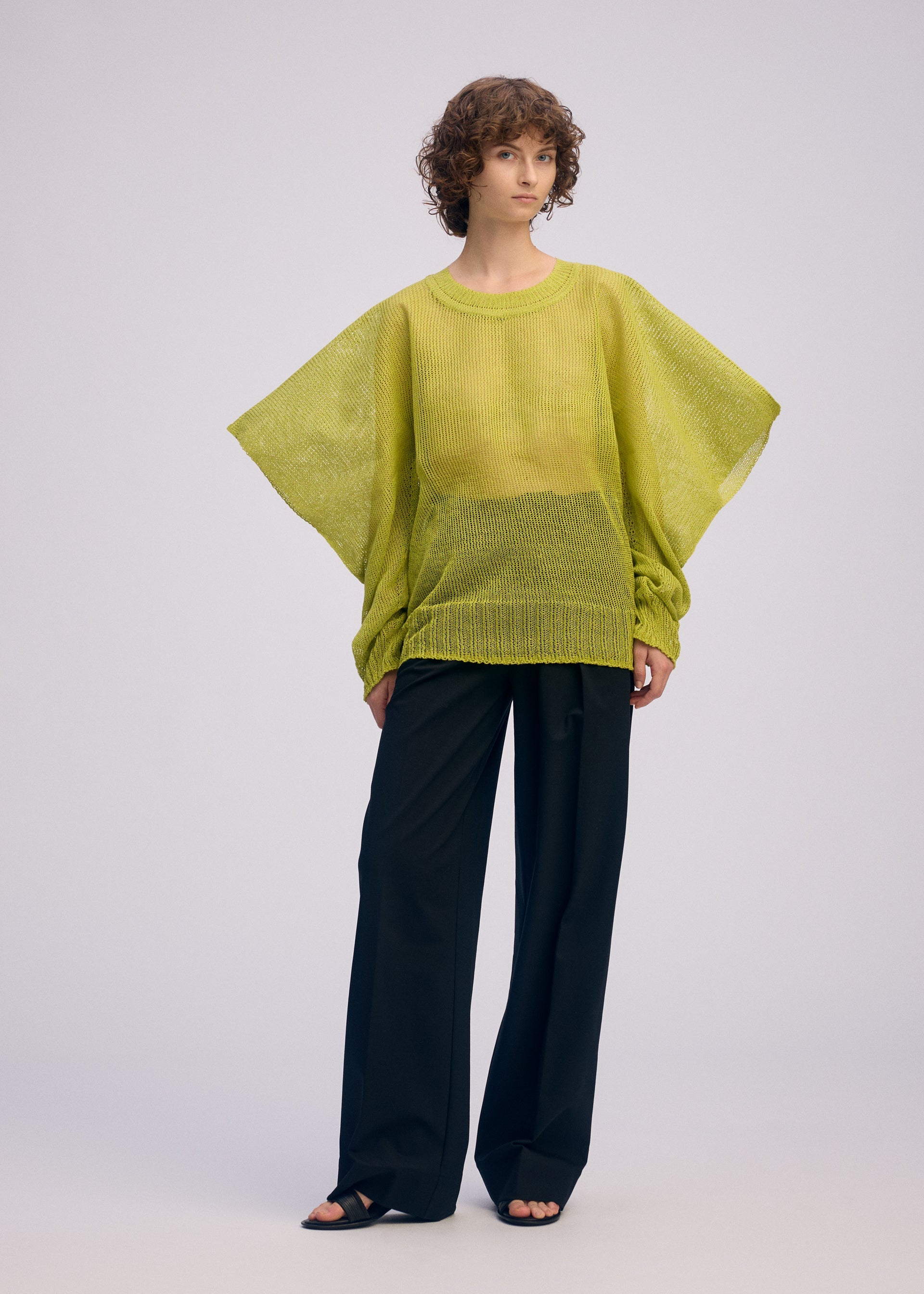 Zoomed view A person with short curly hair wears the oversized lime green CIALDA mesh-knit top by ISSEY MIYAKE with wide sleeves, black wide-leg pants, and sandals, creating a voluminous silhouette against a plain light background.