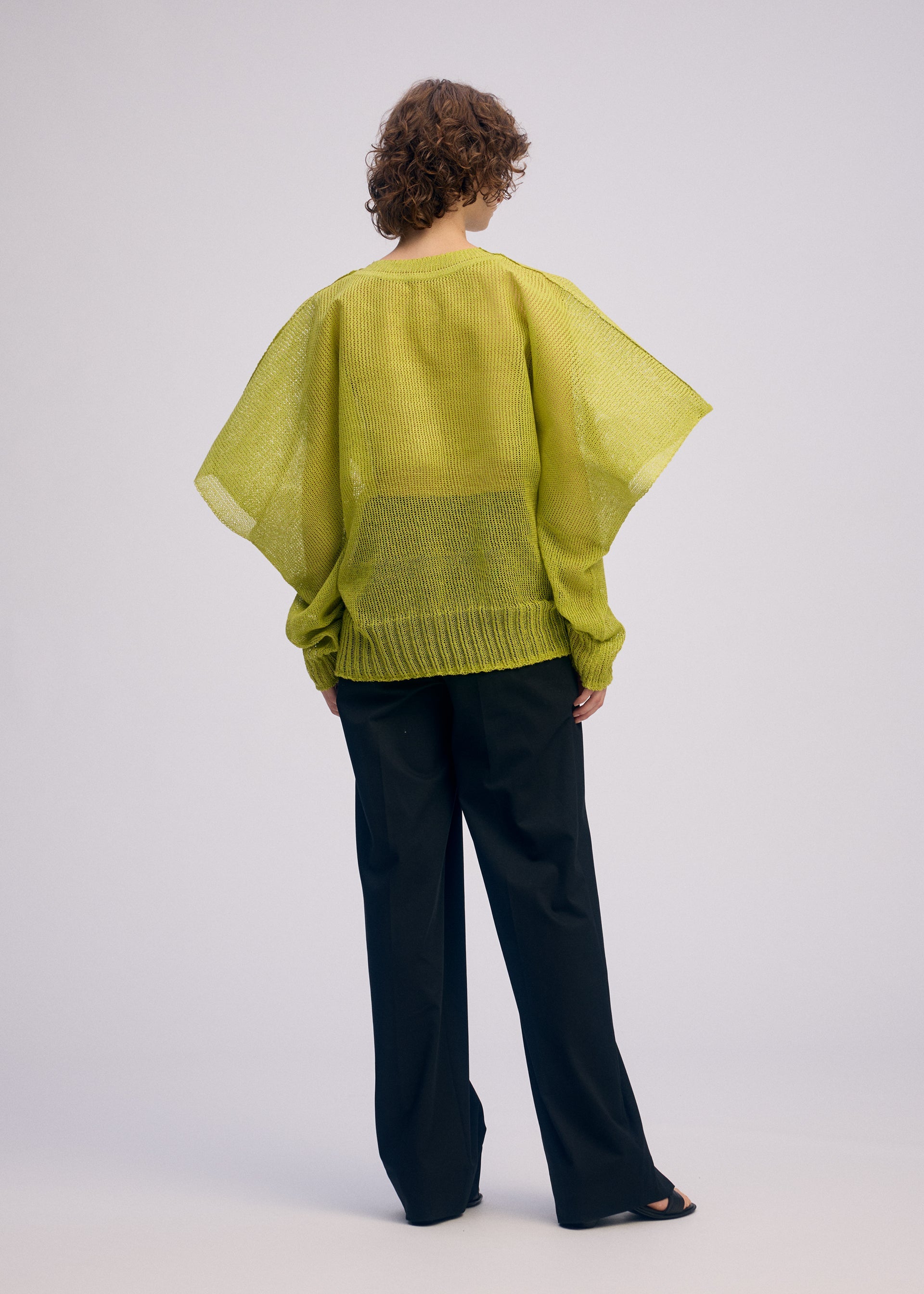 Zoomed view A person with curly hair is seen from behind, wearing the sheer lime green CIALDA mesh-knit top with wide sleeves and loose black trousers by ISSEY MIYAKE, creating a voluminous silhouette against a plain light background.