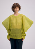 A person with curly brown hair wears the light green CIALDA mesh-like knit top with wide sleeves by ISSEY MIYAKE and black pants, standing against a plain light background.