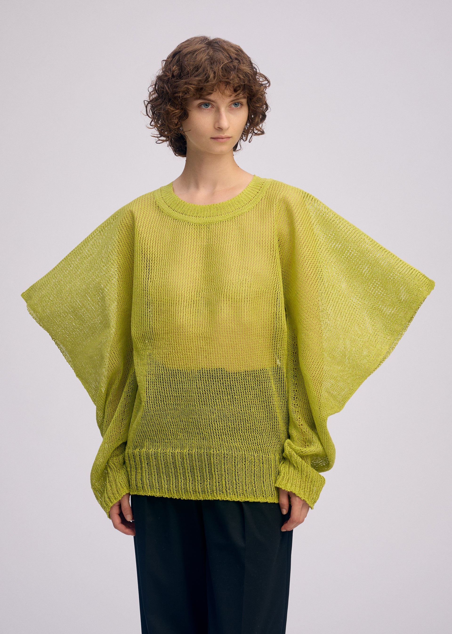 Zoomed view A person with curly brown hair wears the light green CIALDA mesh-like knit top with wide sleeves by ISSEY MIYAKE and black pants, standing against a plain light background.