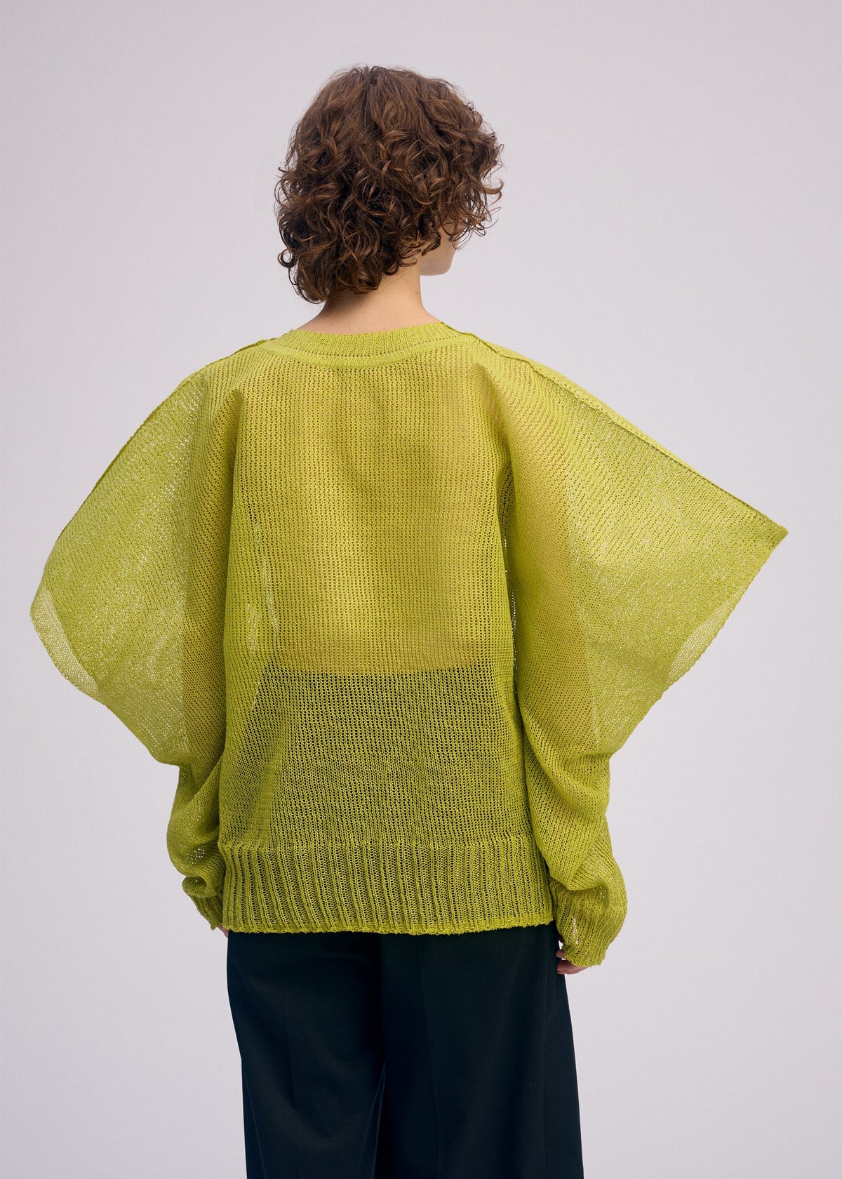 A person with short, curly brown hair is shown from the back wearing the ISSEY MIYAKE CIALDA loose lime green mesh-knit top with wide sleeves and a voluminous silhouette, set against a plain light background. view 9 of 10