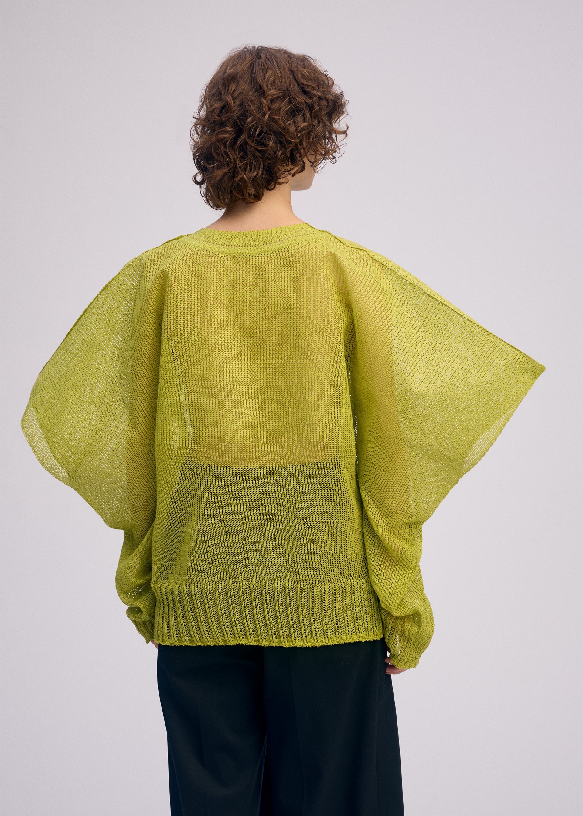 Zoomed view A person with short, curly brown hair is shown from the back wearing the ISSEY MIYAKE CIALDA loose lime green mesh-knit top with wide sleeves and a voluminous silhouette, set against a plain light background.