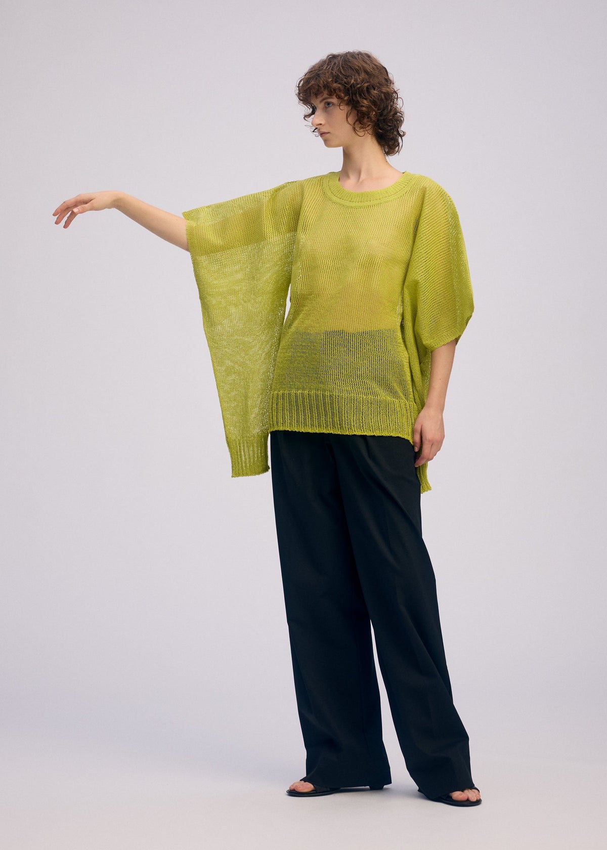 A person with curly hair models the ISSEY MIYAKE CIALDA—a mesh-like knit lime green top—styled with wide-leg black pants, standing sideways with one arm extended against a plain light background. view 10 of 10