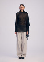 A person faces forward against a plain light background, wearing the ISSEY MIYAKE CONCEALED KNIT top over a black undershirt, paired with loose light gray pants and black sandals.