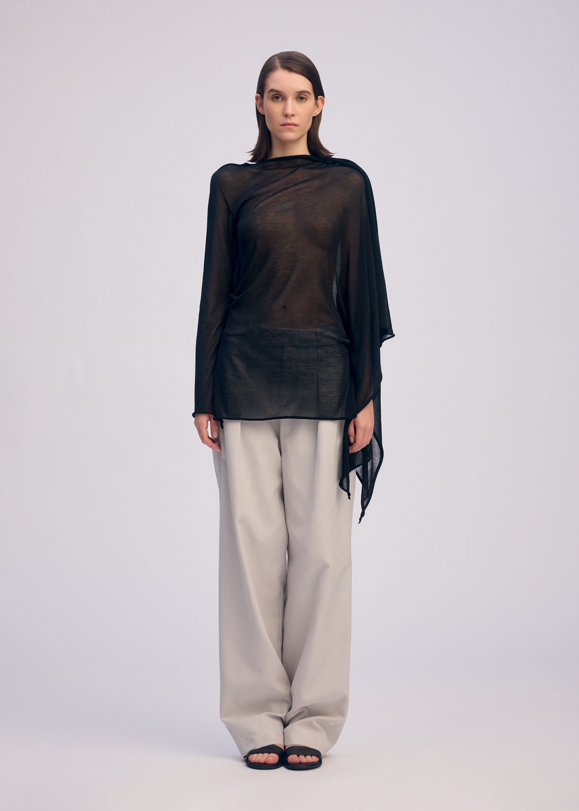 Zoomed view A person faces forward against a plain light background, wearing the ISSEY MIYAKE CONCEALED KNIT top over a black undershirt, paired with loose light gray pants and black sandals.